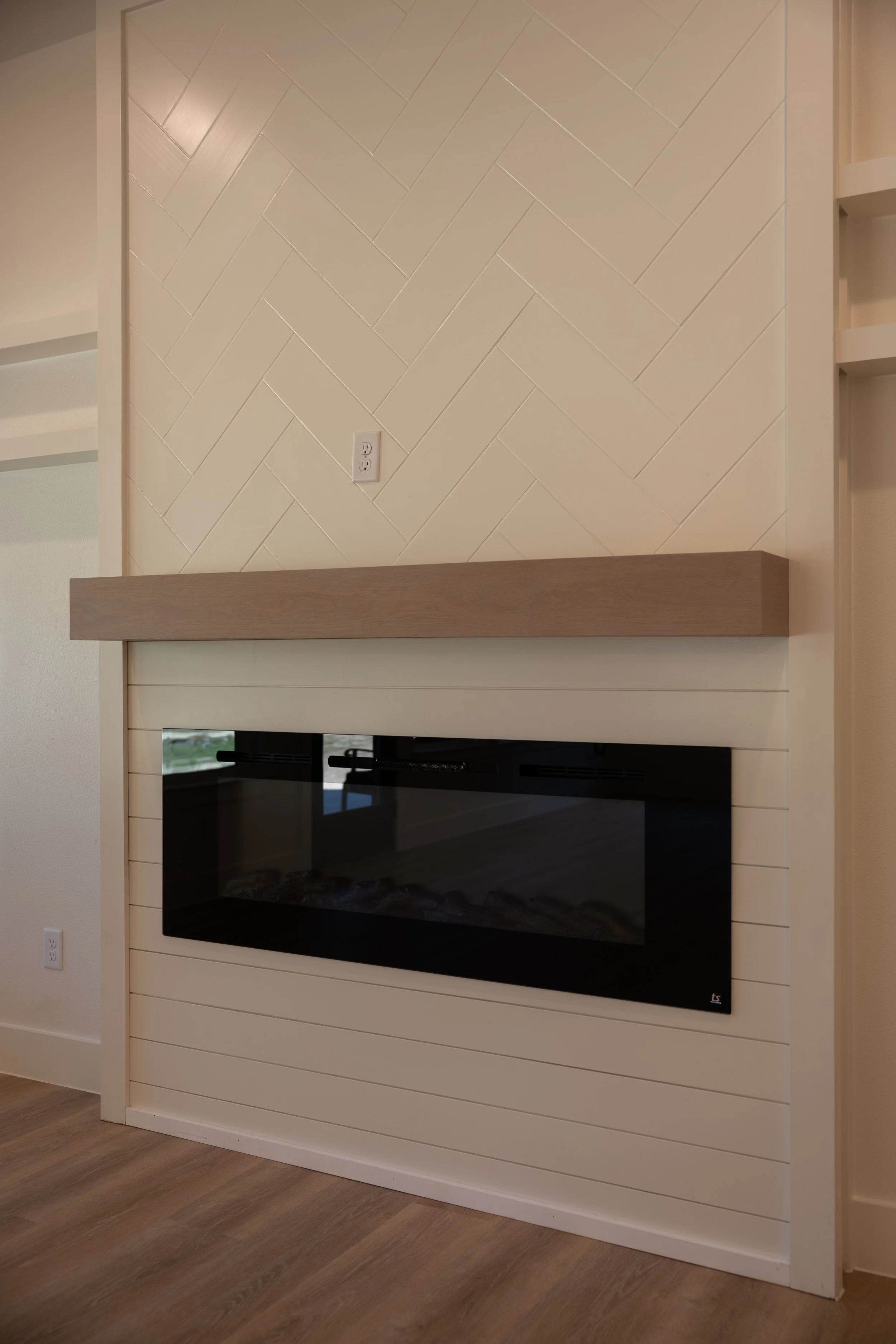 A modern living room fireplace with a built-in electric or wood-burning fireplace with a black glass front, surrounded by white horizontal wood paneling. Above the fireplace is a beige wall with a diagonal tile pattern, featuring a white electrical o