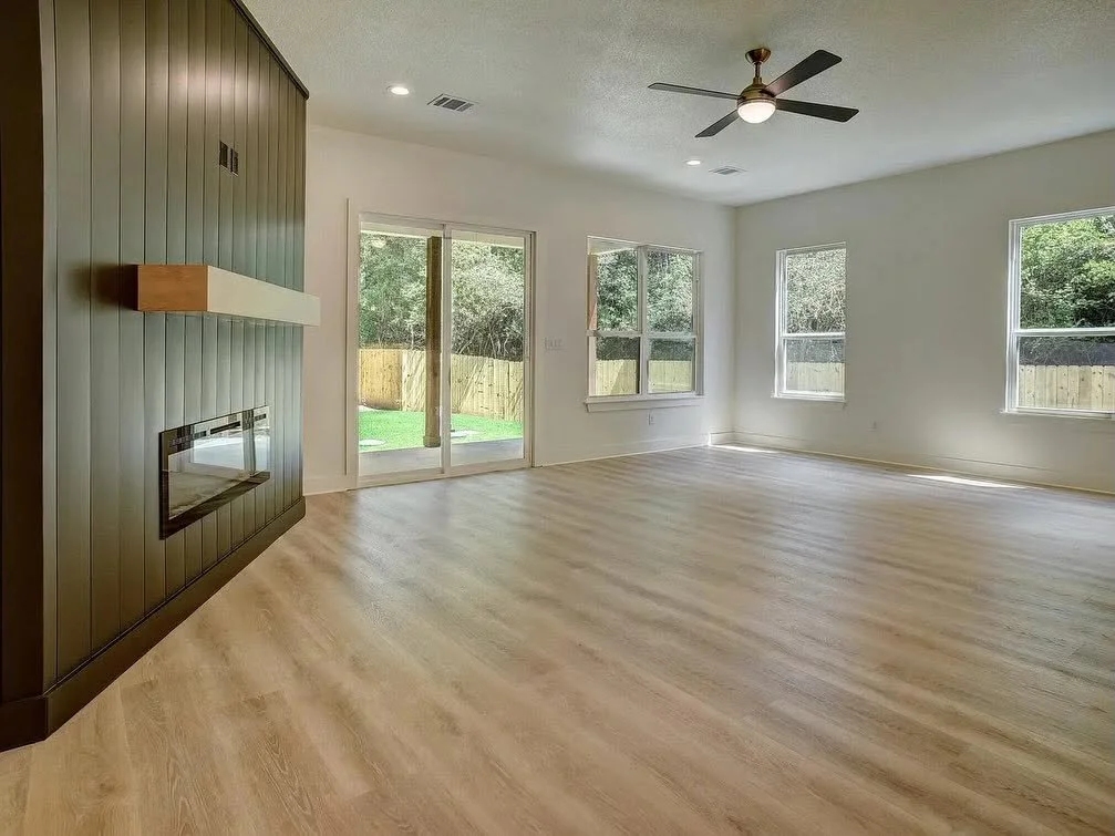 Empty living room with light wood flooring, white walls, a ceiling fan, large windows, and a sliding glass door leading to the backyard with a fence and trees.