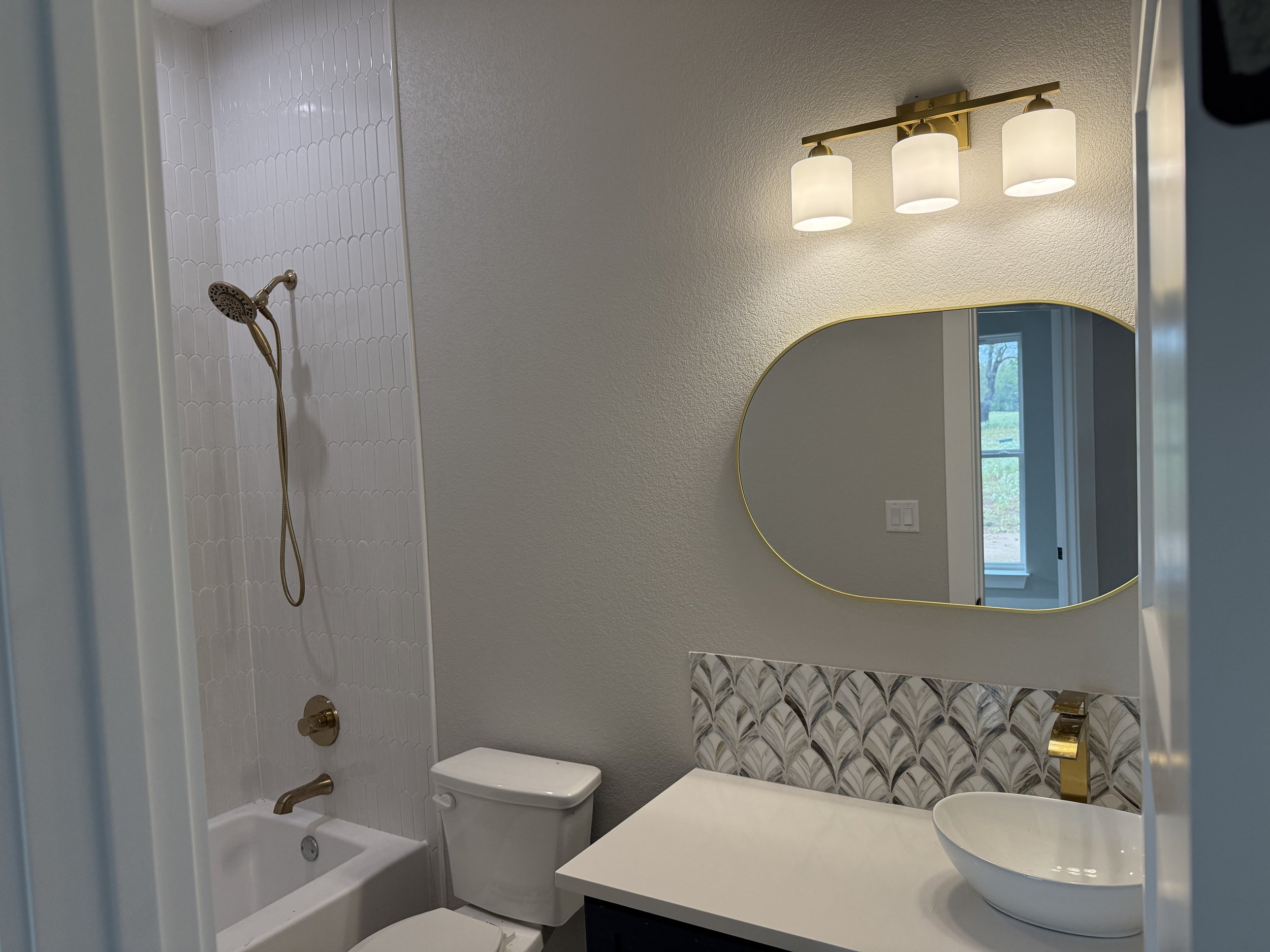 Modern bathroom with a walk-in shower, toilet, and vanity with vessel sink. The shower features white tile with a gold showerhead, and there is a round mirror above the vanity. The lighting fixture has three white glass shades, and a window showing outdoor scenery is visible in the mirror reflection.