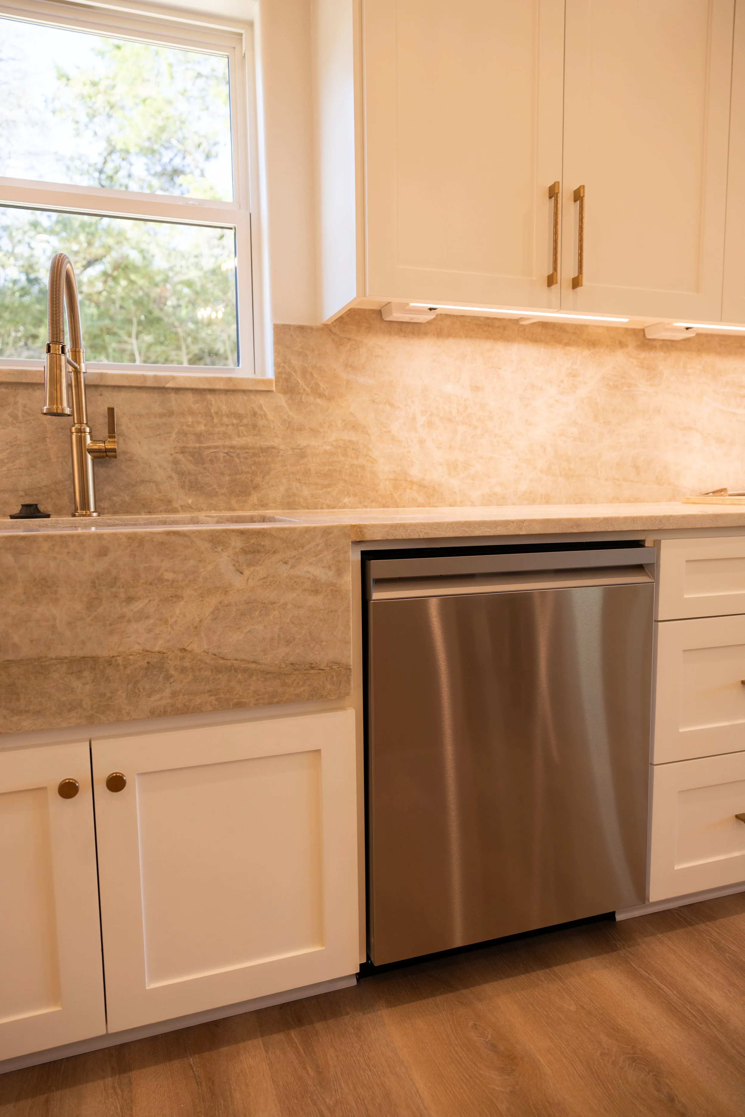A kitchen with beige countertop and backsplash, white cabinets with gold handles, a stainless steel dishwasher, a double-hung window showing trees outside, and a stainless steel faucet over the sink.