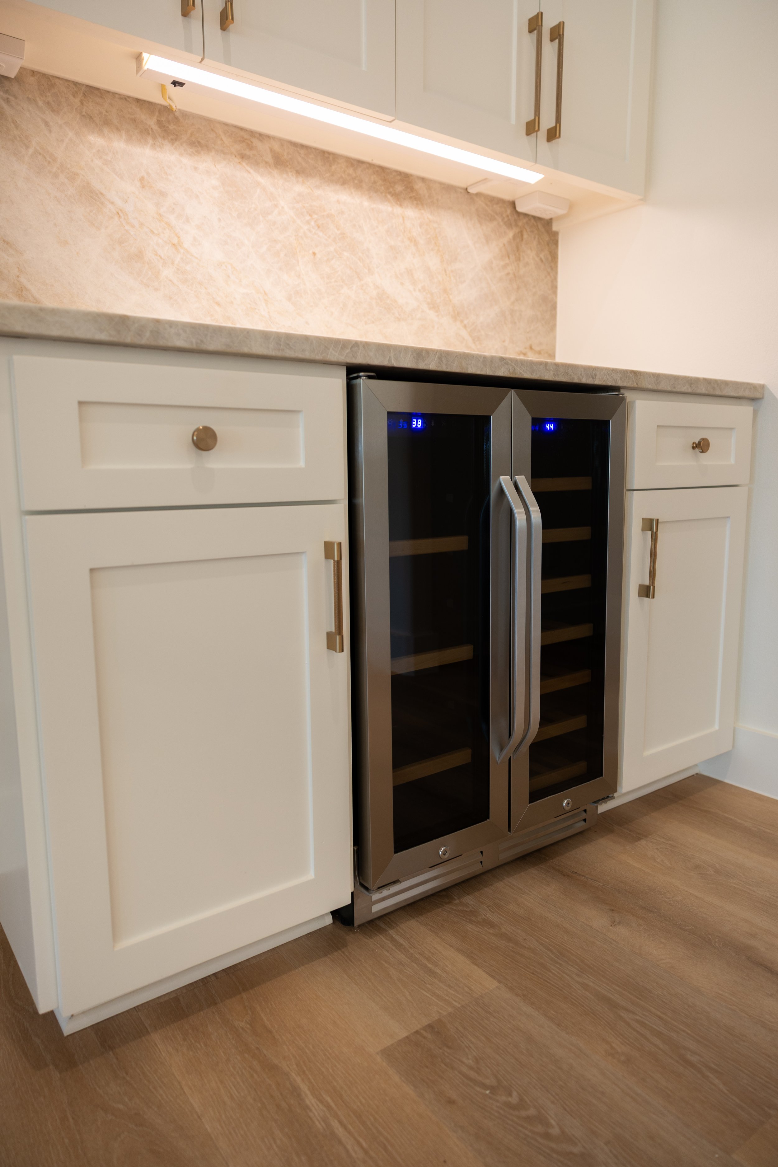 A modern kitchen with white cabinets, a marble countertop, and a built-in glass-front wine cooler. The floor is wood, and there is under-cabinet lighting.