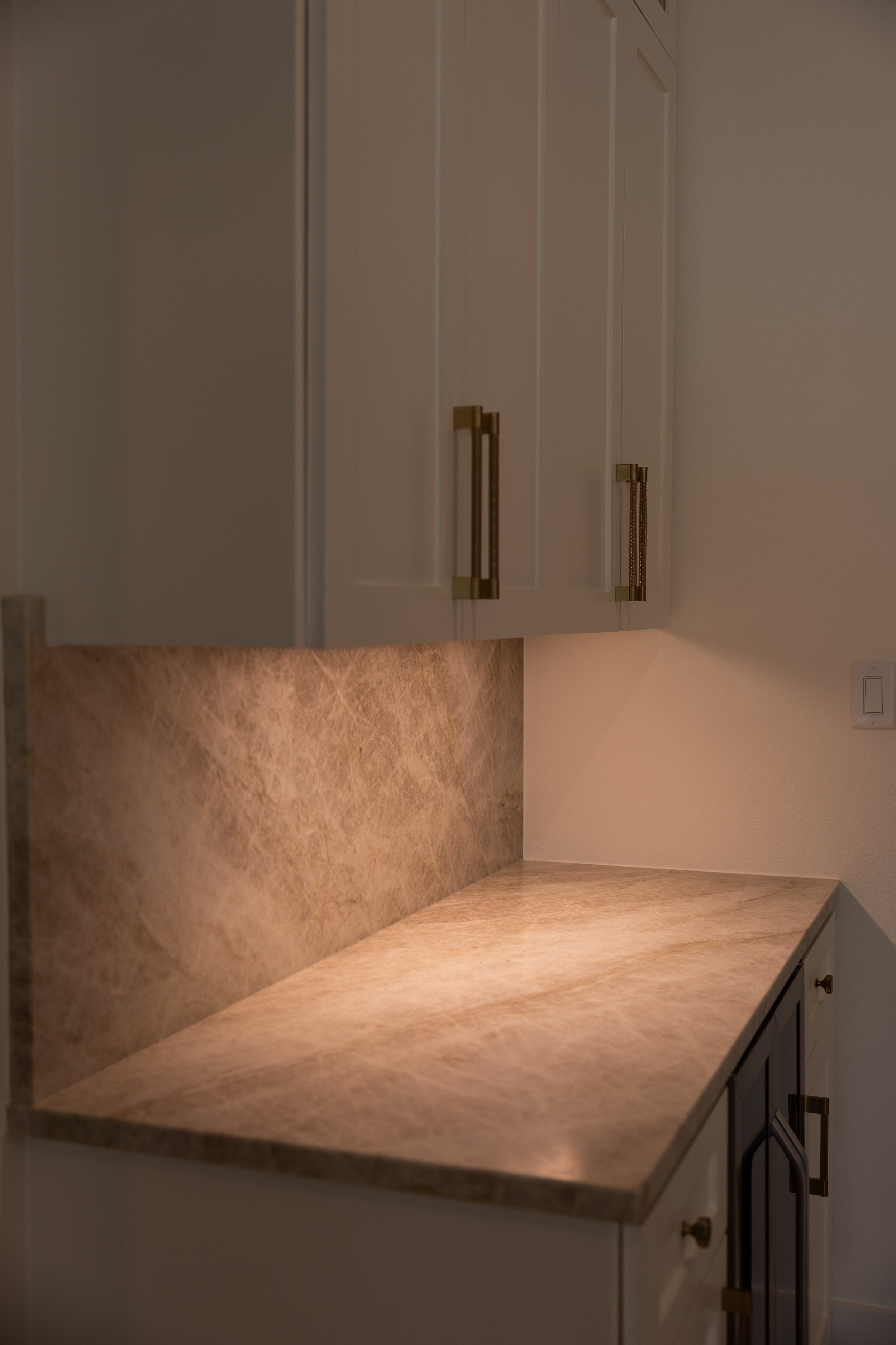 Kitchen countertop with beige marble pattern, white cabinets with brass handles, beige backsplash, and a white wall with a light switch.