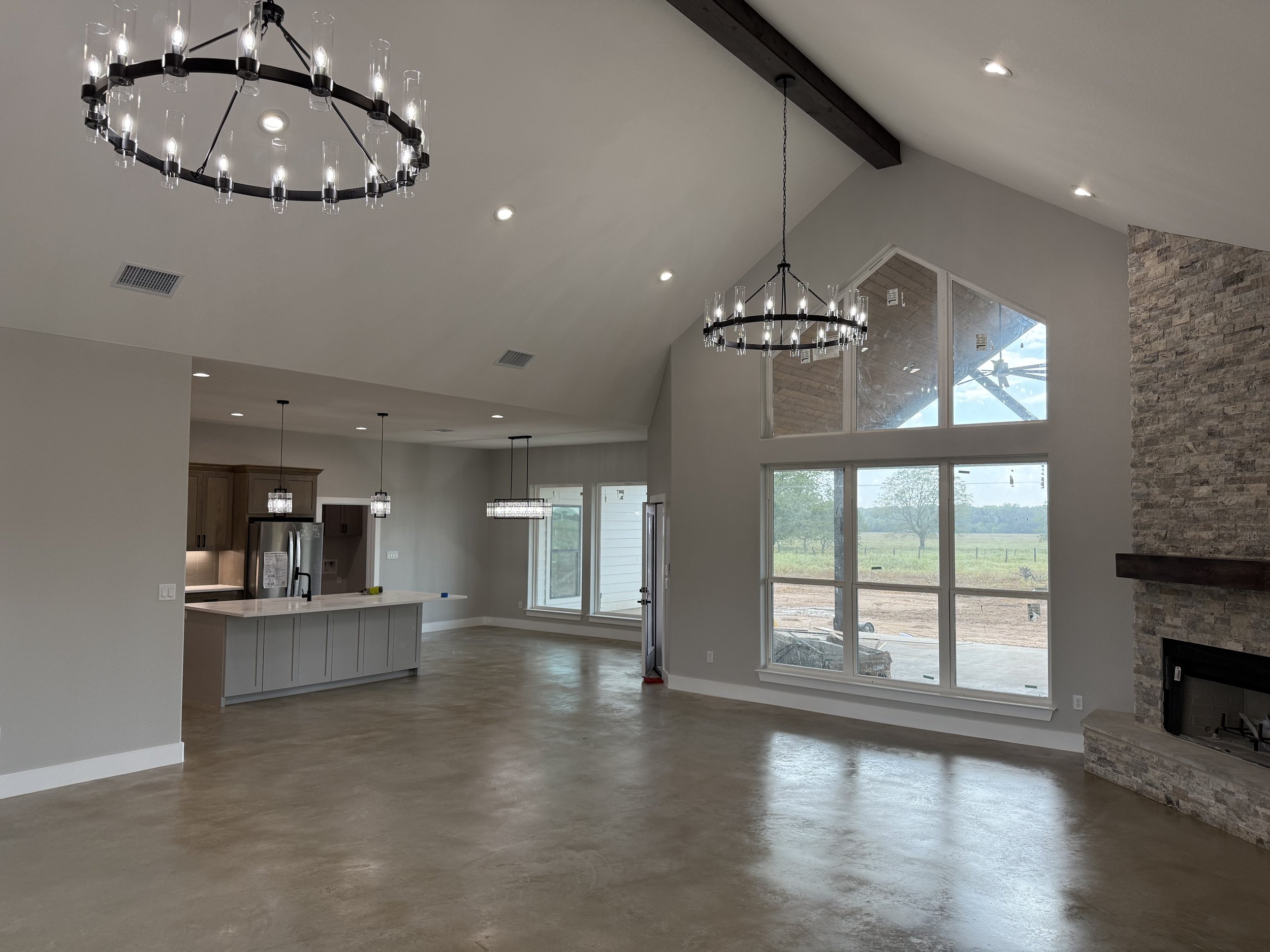 Empty living room with large window, stone fireplace, high vaulted ceiling, and modern chandelier lights.