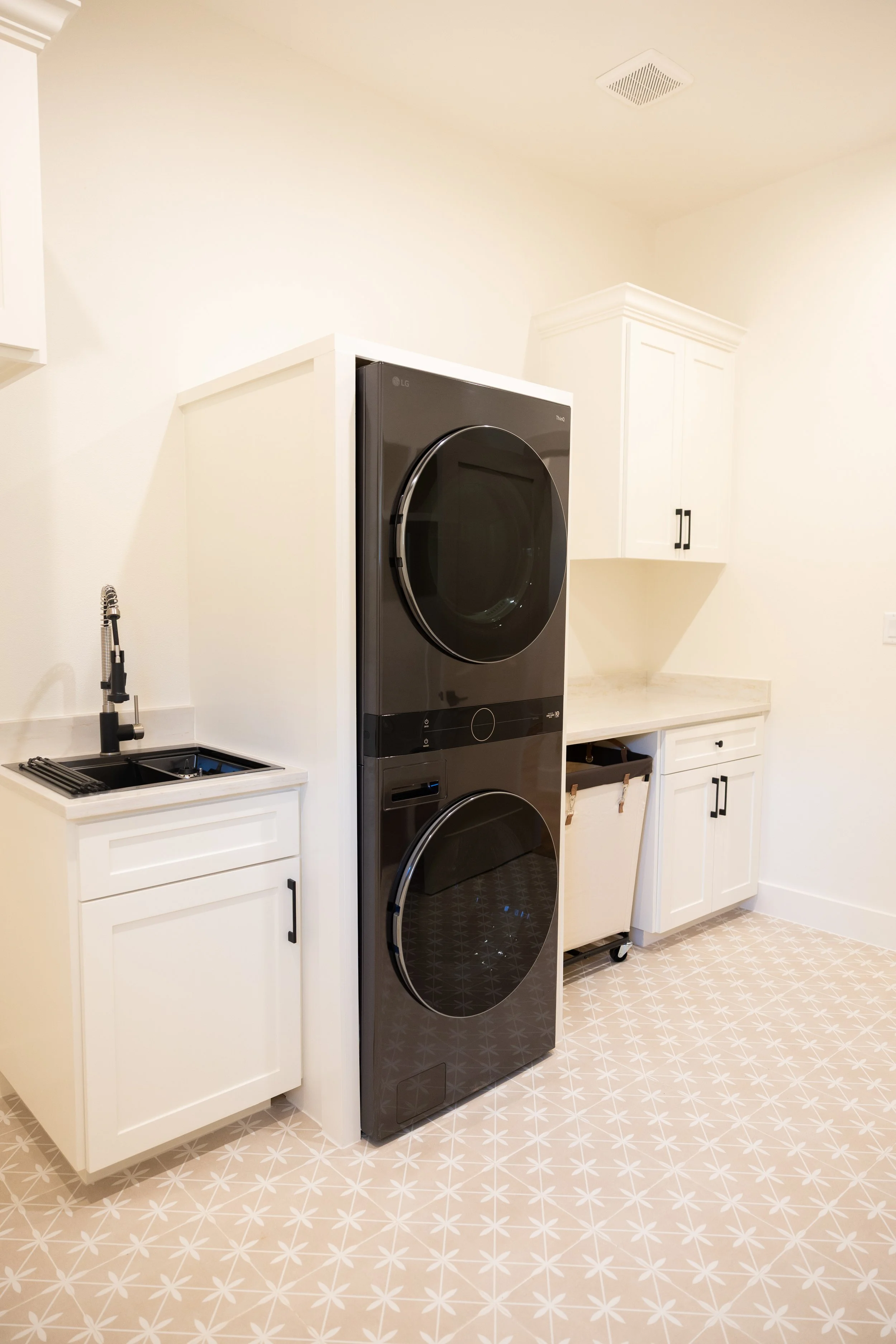 A laundry room with white cabinets, a black LG washer and dryer stacked, a small sink and faucet, and patterned tile flooring.