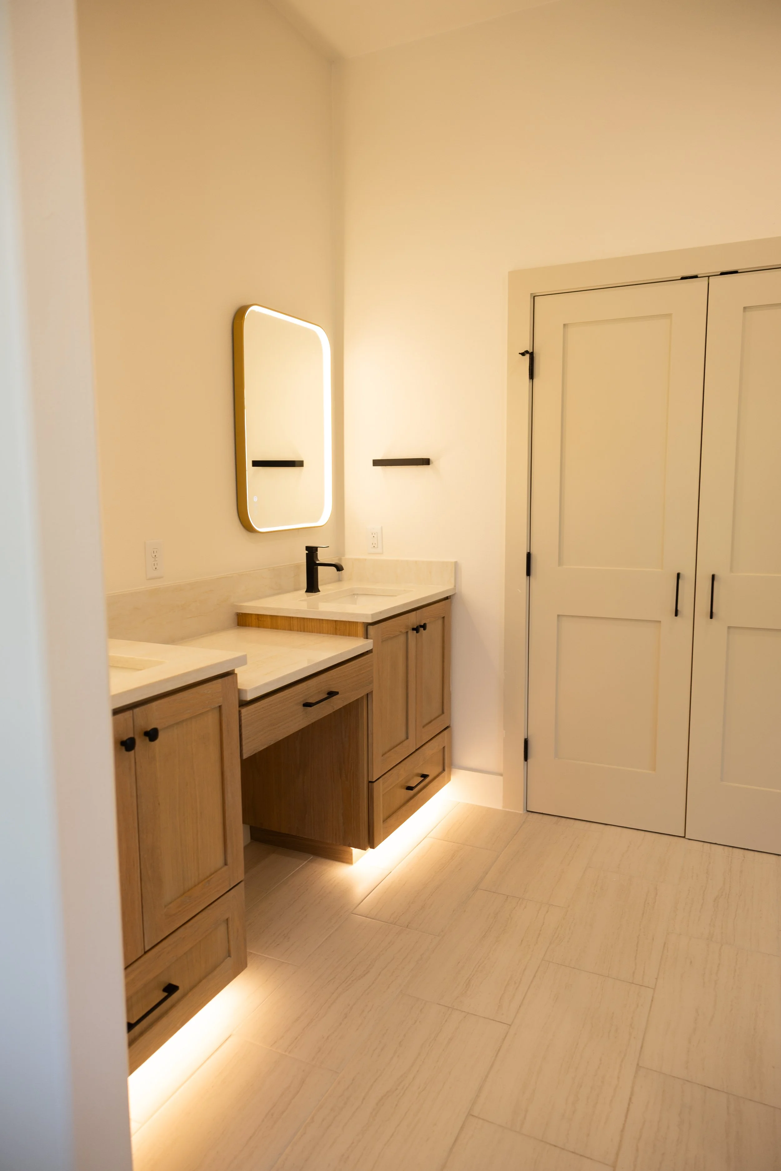 A modern bathroom with light wood cabinets, a white marble countertop, a black faucet, a backlit mirror, and a closed white closet door with black hinges.