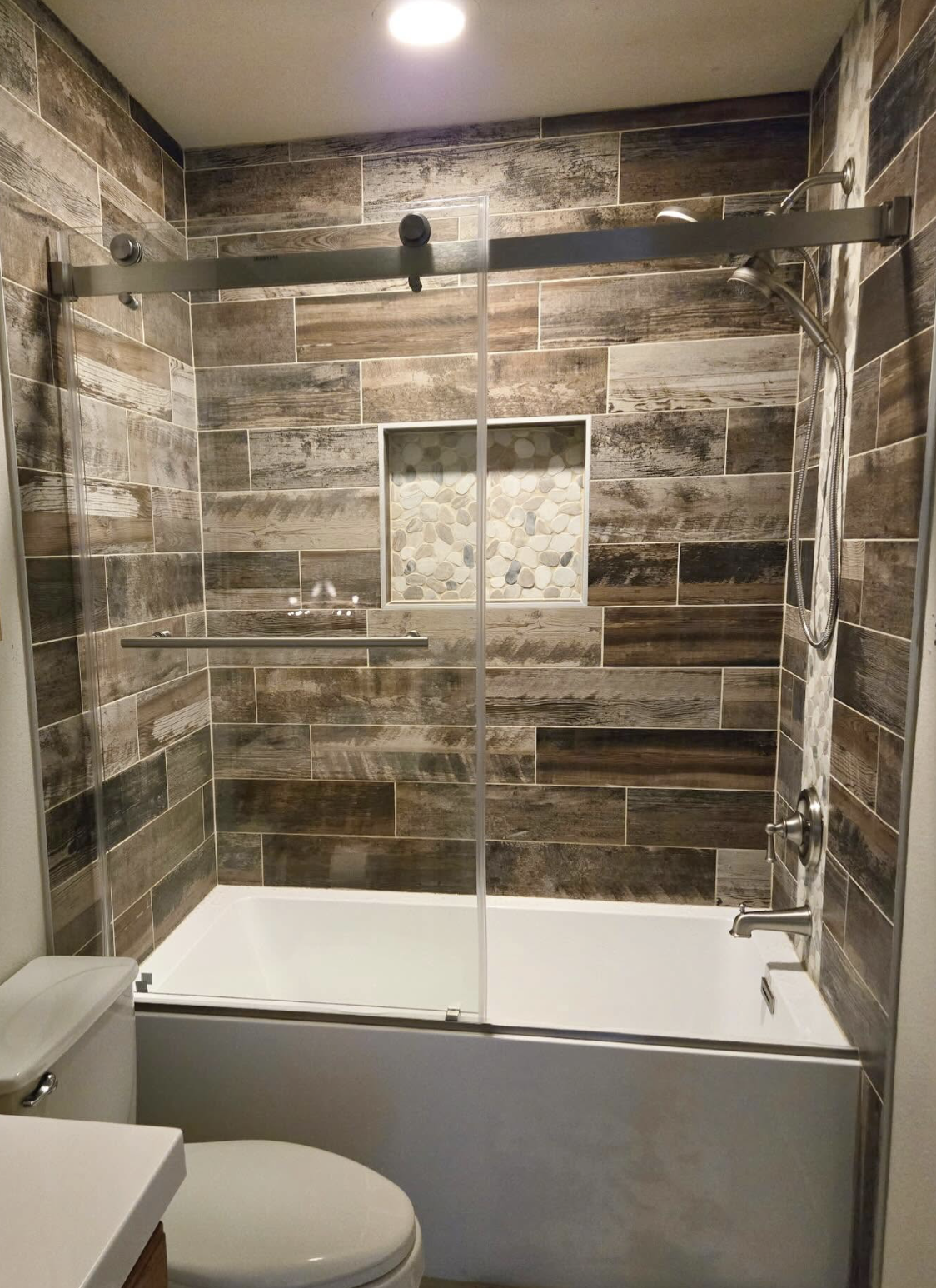 Bathroom shower with wood-look tile walls, a built-in pebble niche, glass sliding door, and a handheld showerhead.