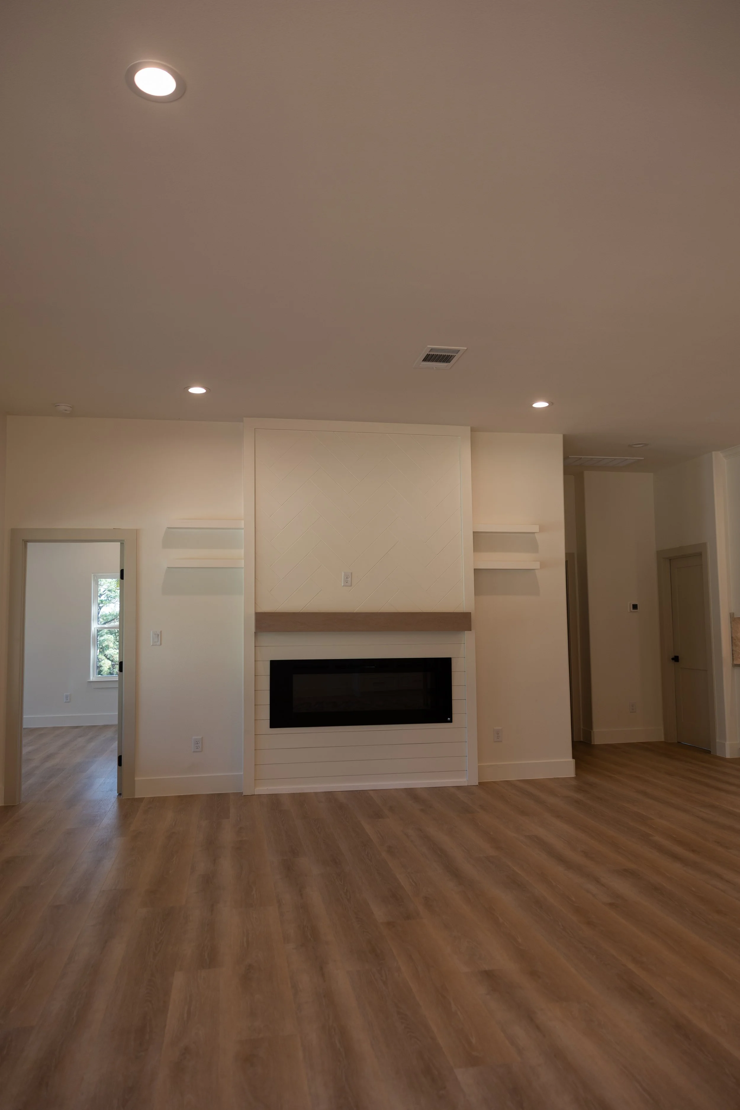 Interior view of a living room with wood flooring, a fireplace, cream-colored walls, a small window, and ceiling lights.