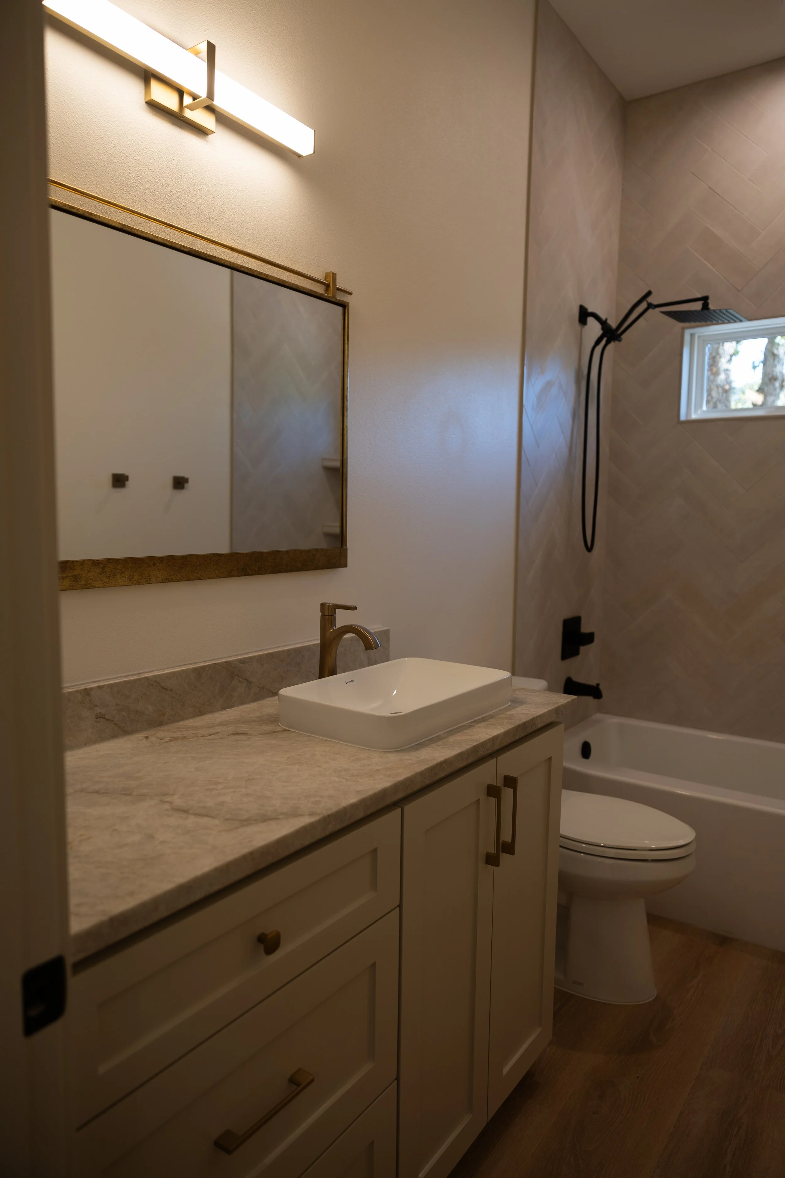 Bathroom with beige vanity, square vessel sink, gold hardware, wall-mounted mirror, wall light, and a shower with a black rain shower head and a small window.