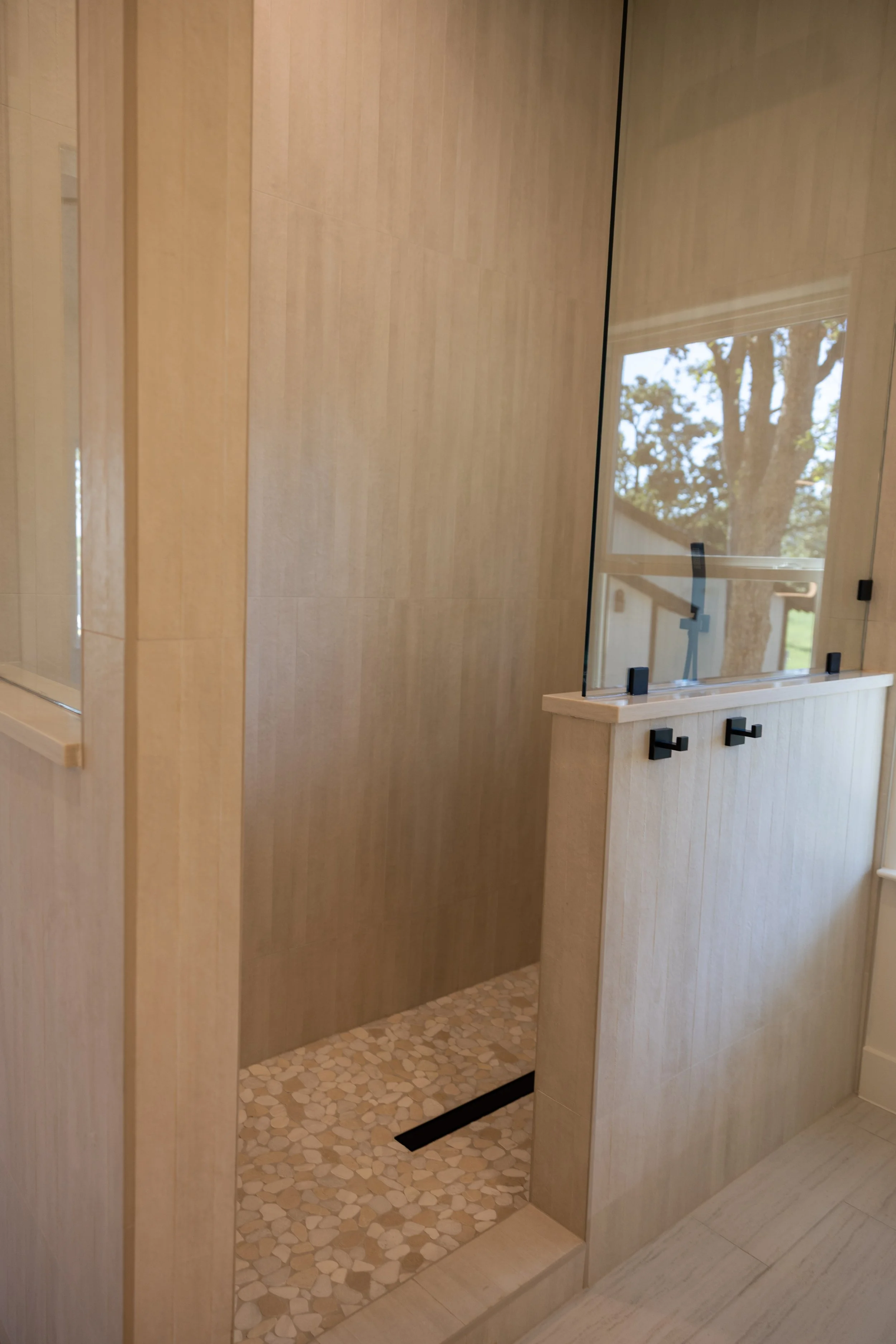 Bathroom with a walk-in shower, natural wood paneling, a window showing trees outside, and a small built-in shelf with black hooks.
