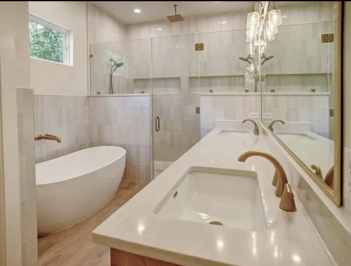Modern bathroom with a white freestanding bathtub, glass-enclosed shower, double vanity with two sinks, and a large mirror with gold fixtures, illuminated by warm lighting.