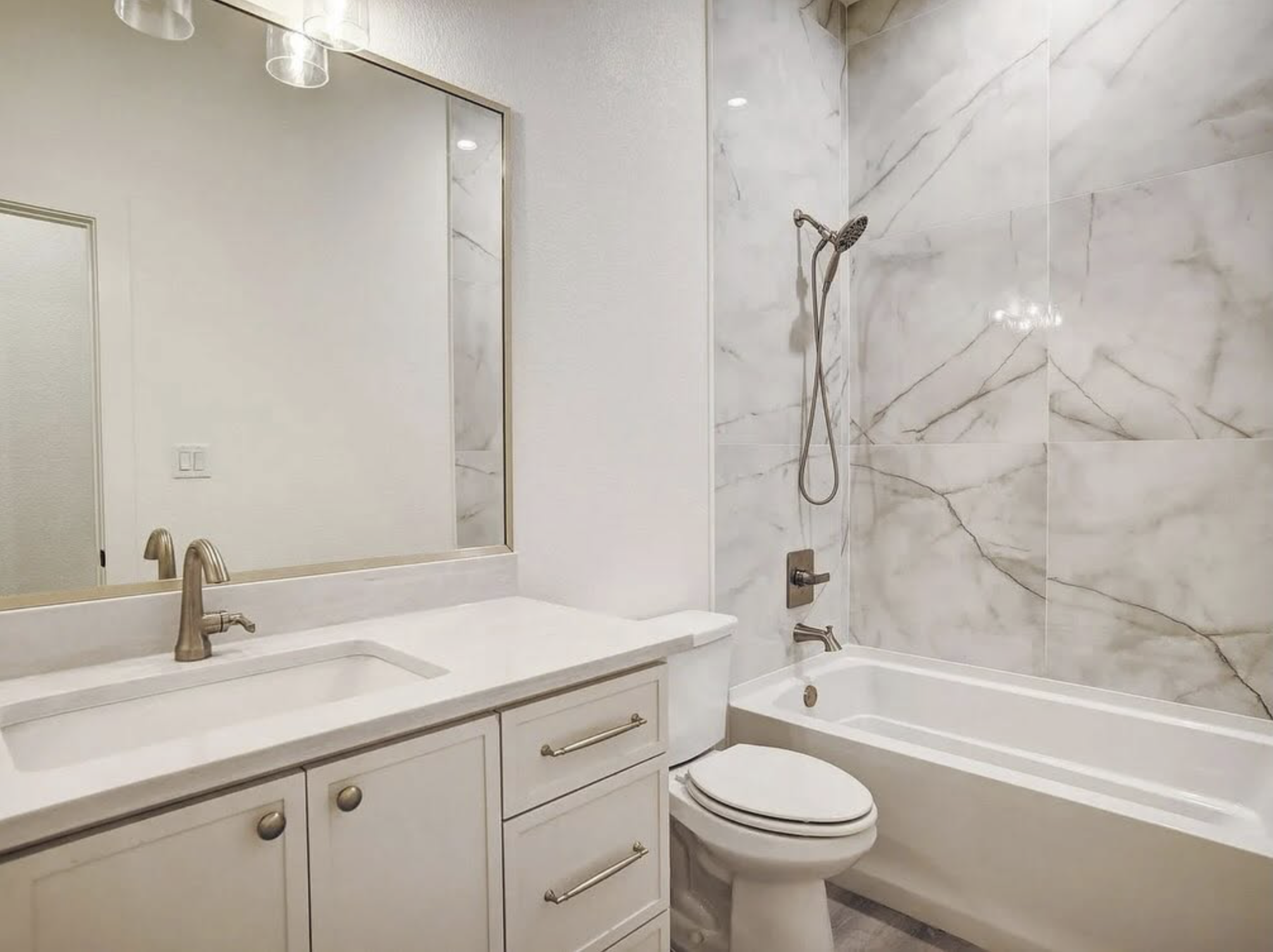 A modern bathroom with a large mirror, marble-tiled walls, a white sink with a gold faucet, and a white bathtub. The bathroom also features a toilet and sleek fixtures.