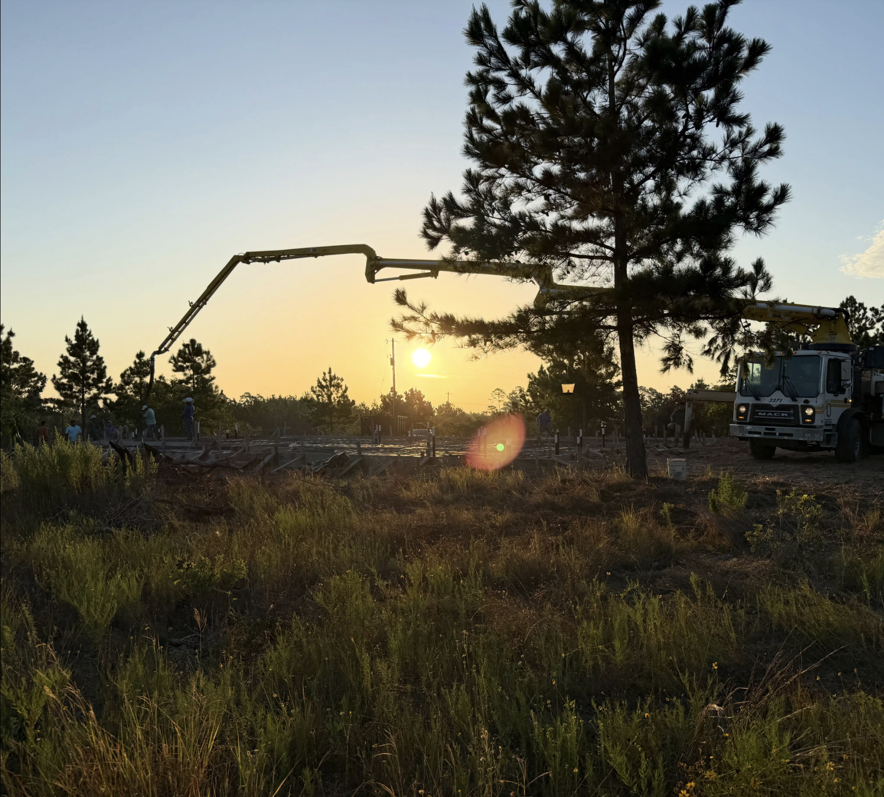 Sunset over a construction site with a crane lifting a tree, workers on site, and lush green grass in the foreground.