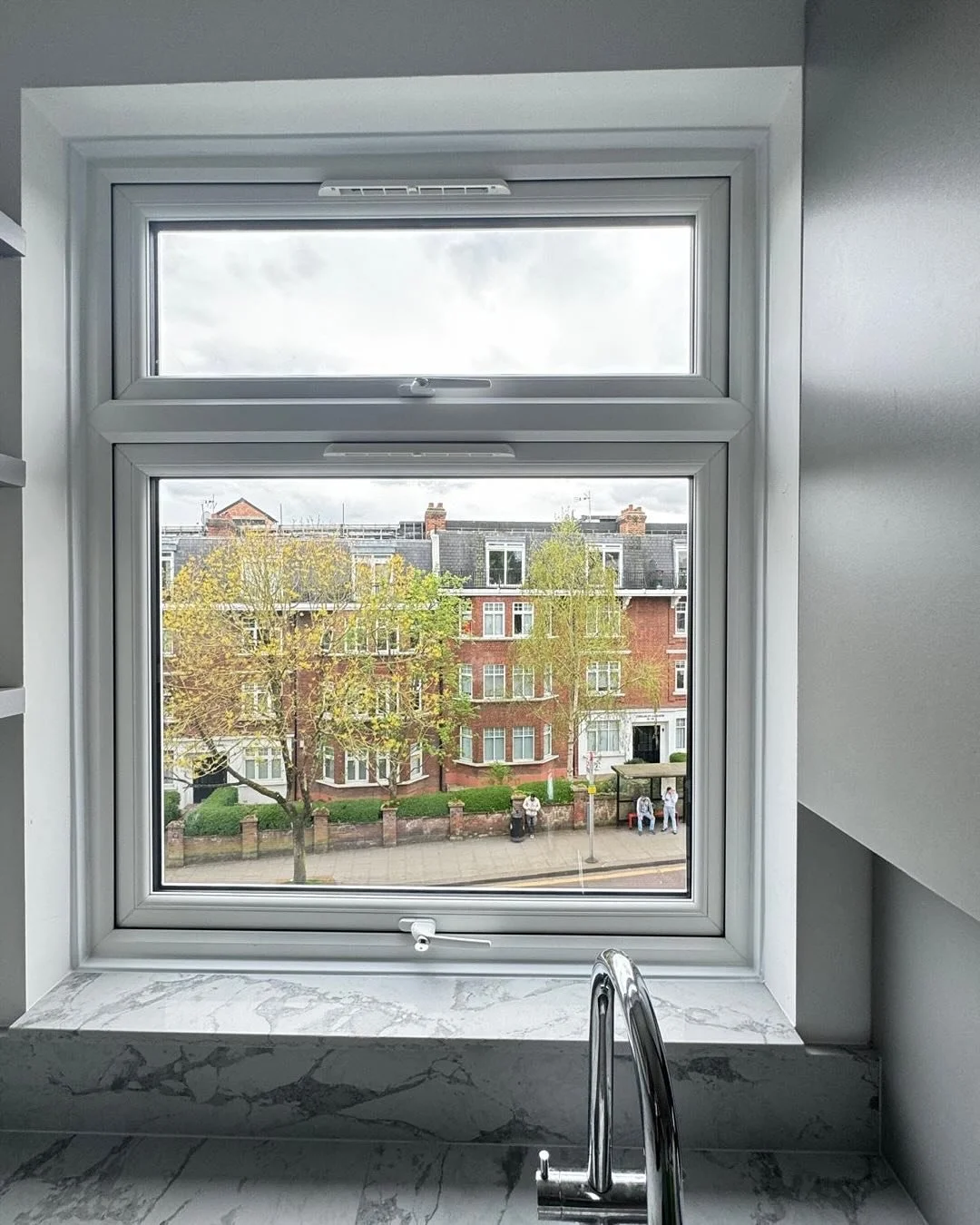View through a bathroom window showing a street with apartment buildings, trees, and two people sitting on a bench.