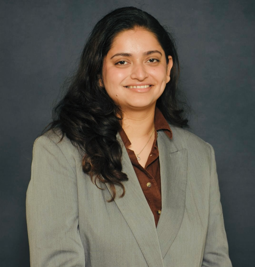 A woman with long black hair and a warm smile wearing a light gray blazer over a brown shirt, standing against a dark background.