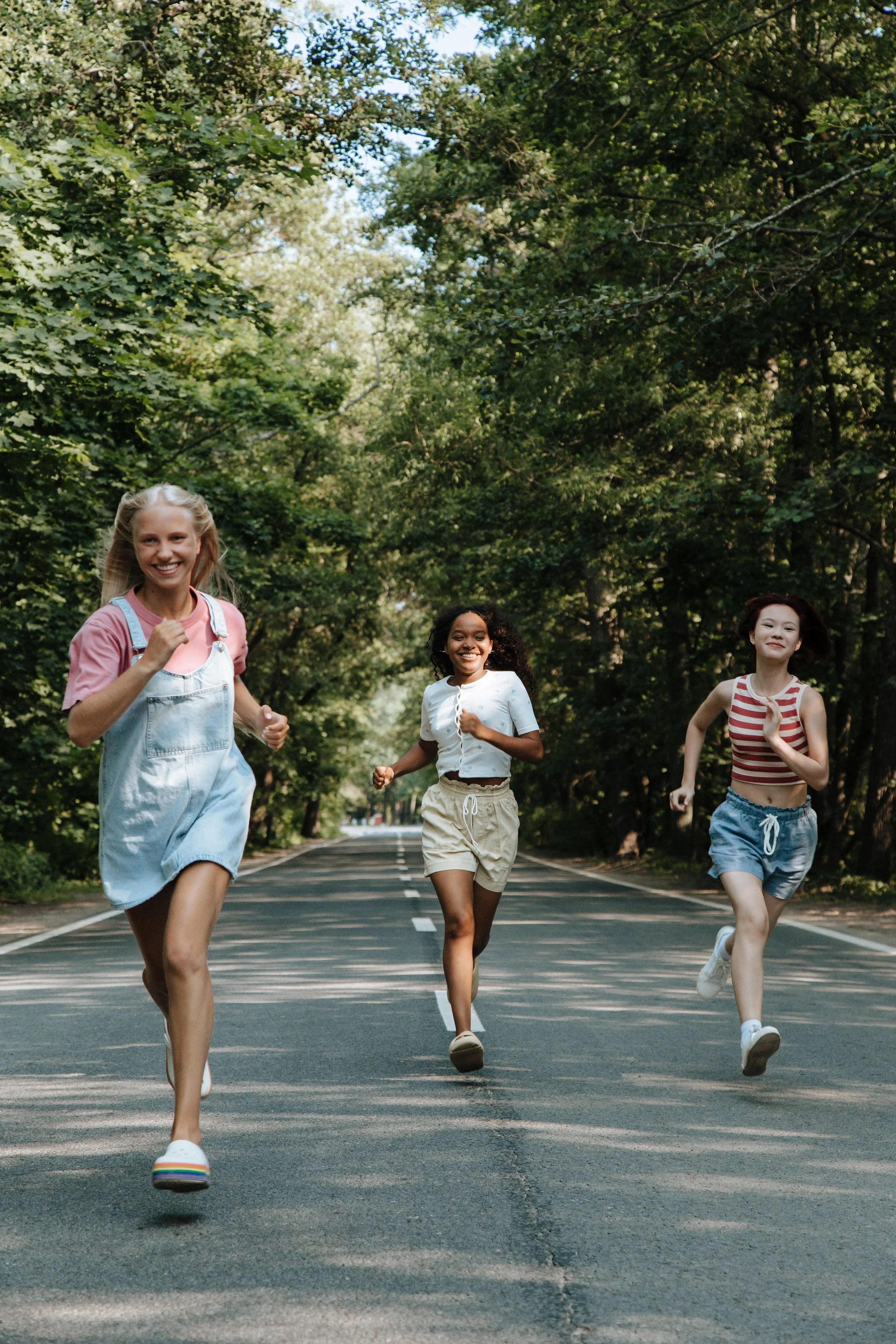 Three young women running on a tree-lined street, smiling and enjoying a sunny day.