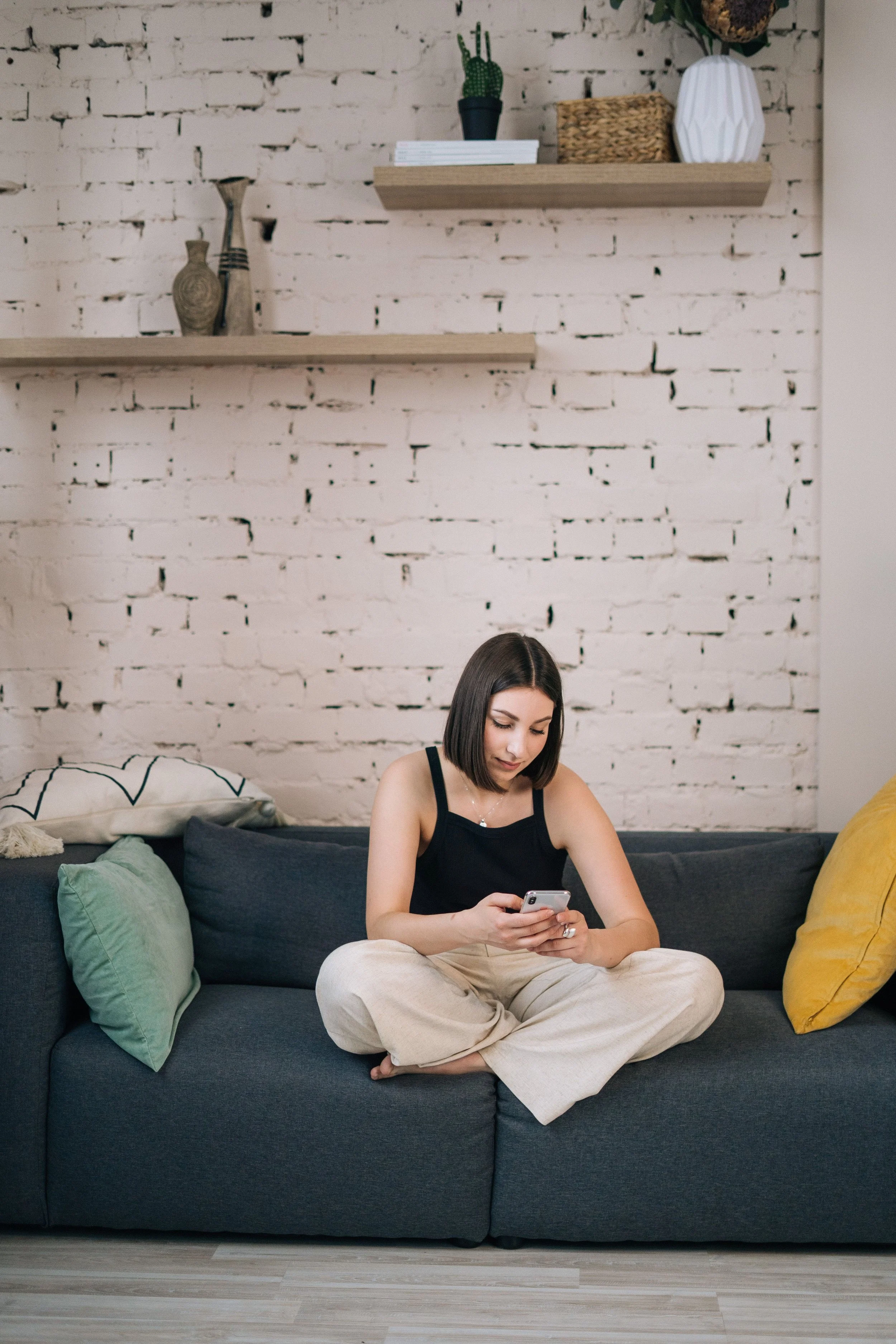 A woman with dark hair, wearing a black top and beige pants, sitting cross-legged on a dark gray sofa, holding and looking at her phone. The sofa has a green pillow on the left and a yellow pillow on the right. There is a white brick wall with floating shelves above her, and a wooden floor.