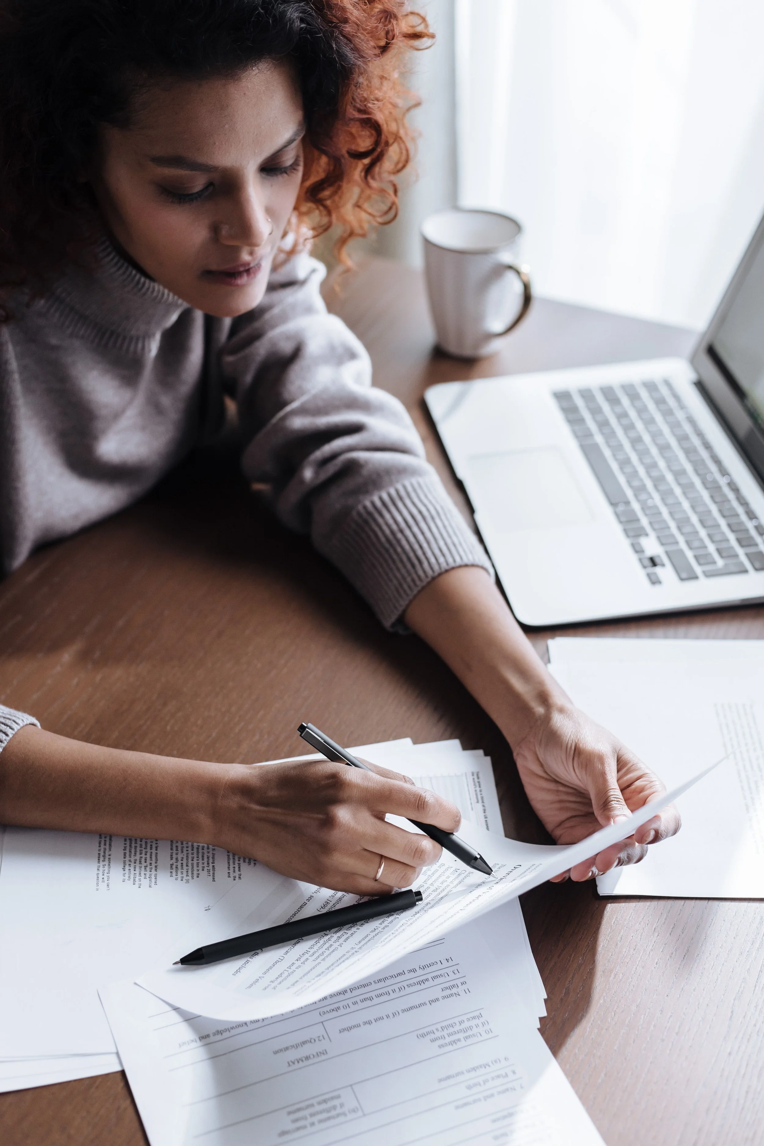 Woman with curly hair working on documents at a desk, surrounded by papers and a laptop, with a coffee mug nearby.