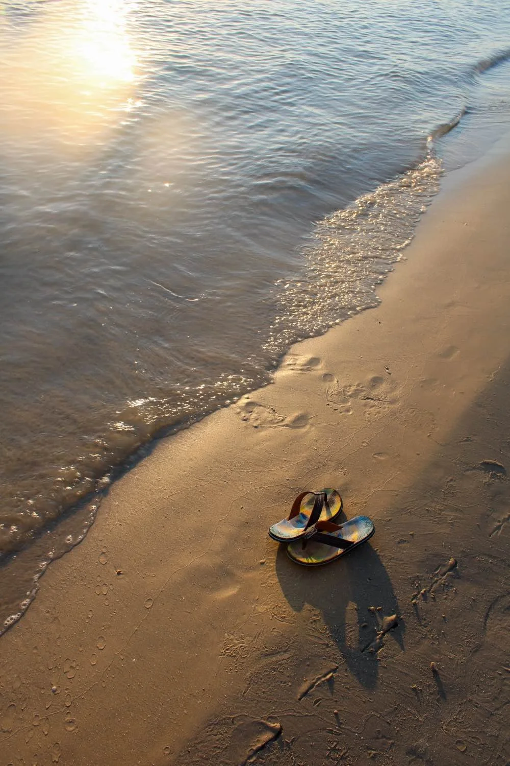 Flip-flops on sandy beach with gentle waves and sunlight reflection on water.