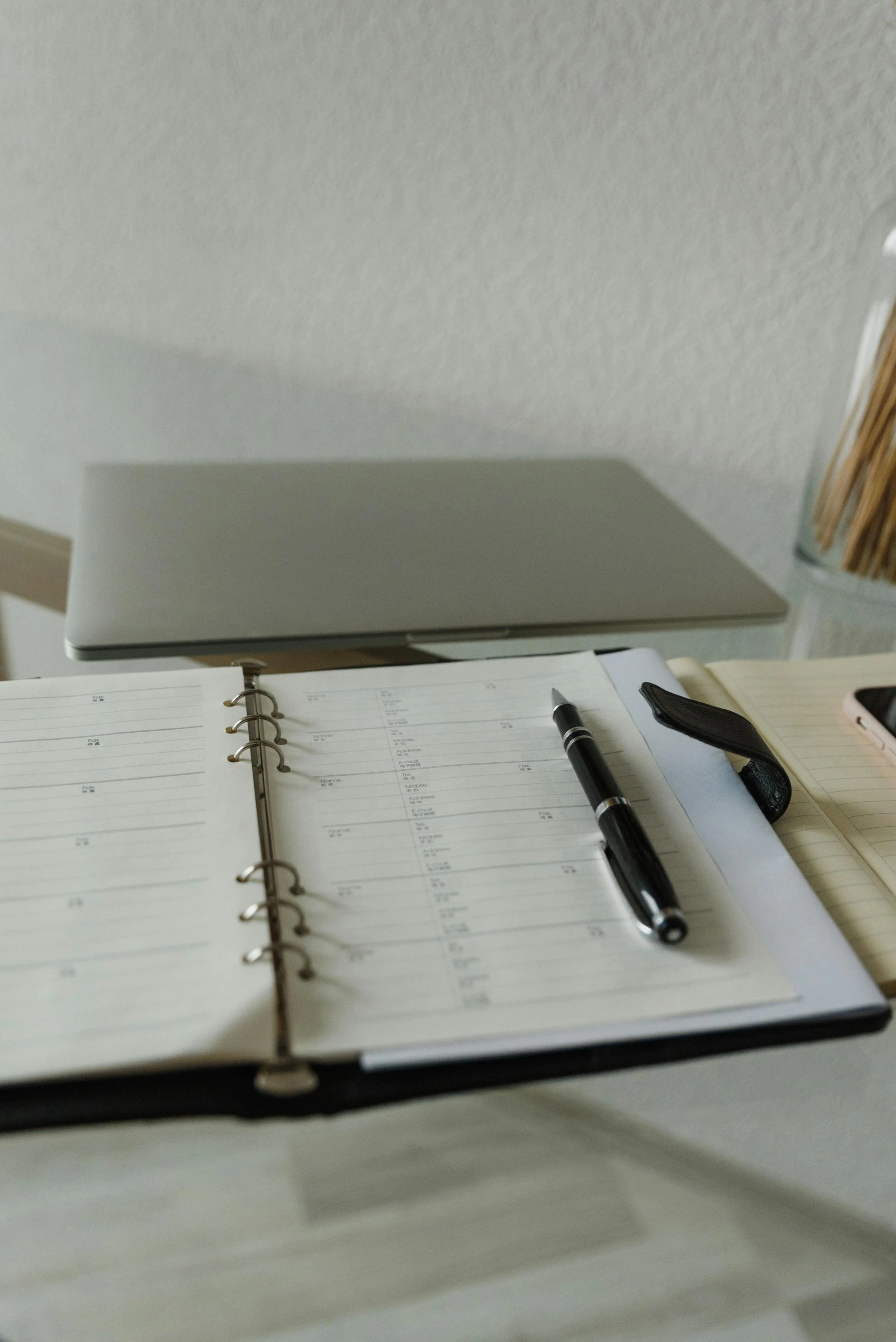 Open planner book with a black pen placed on it, a silver laptop, a set of keys, and a cup of pencils on a desk.