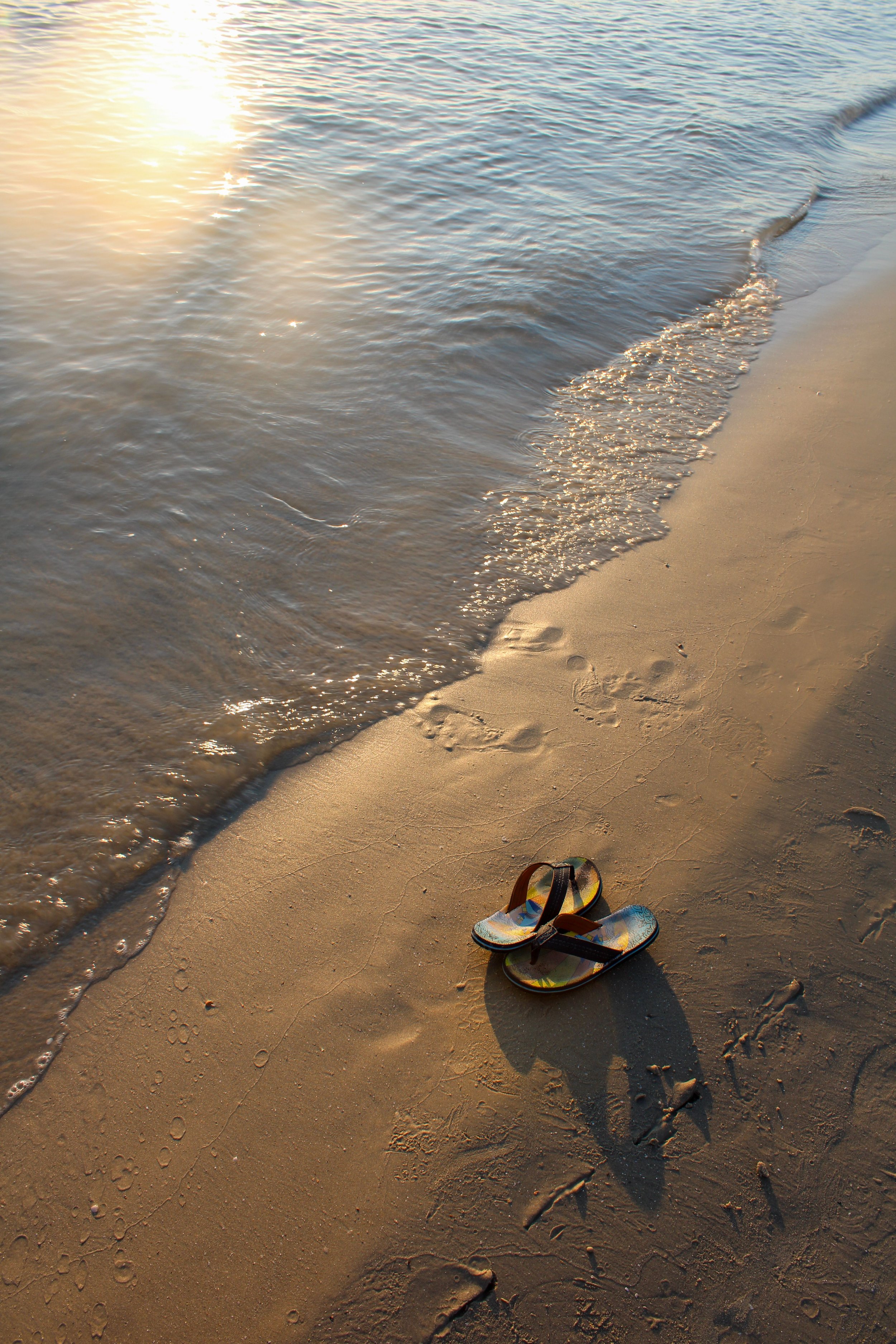 Flip-flops on sandy beach with gentle waves and sunlight reflection on water.