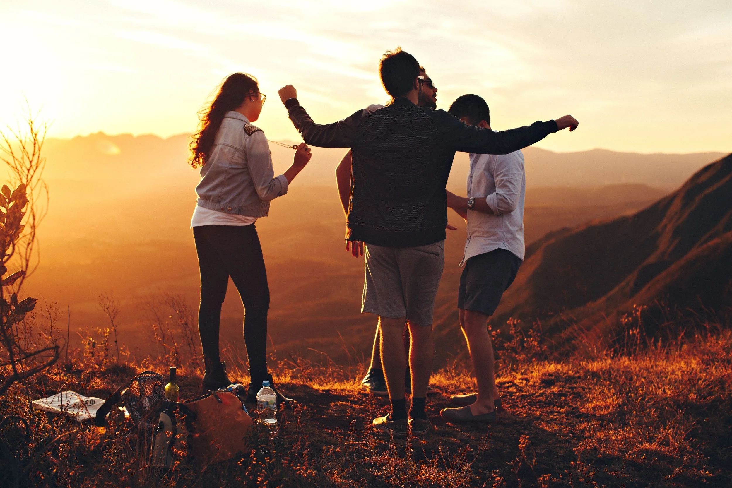 Four friends stand together on a hilltop during sunset, with mountains in the background. They are dressed casually, holding drinks, and appear to be enjoying each other's company. Some items like bags, water bottles, and a blanket are on the ground nearby.