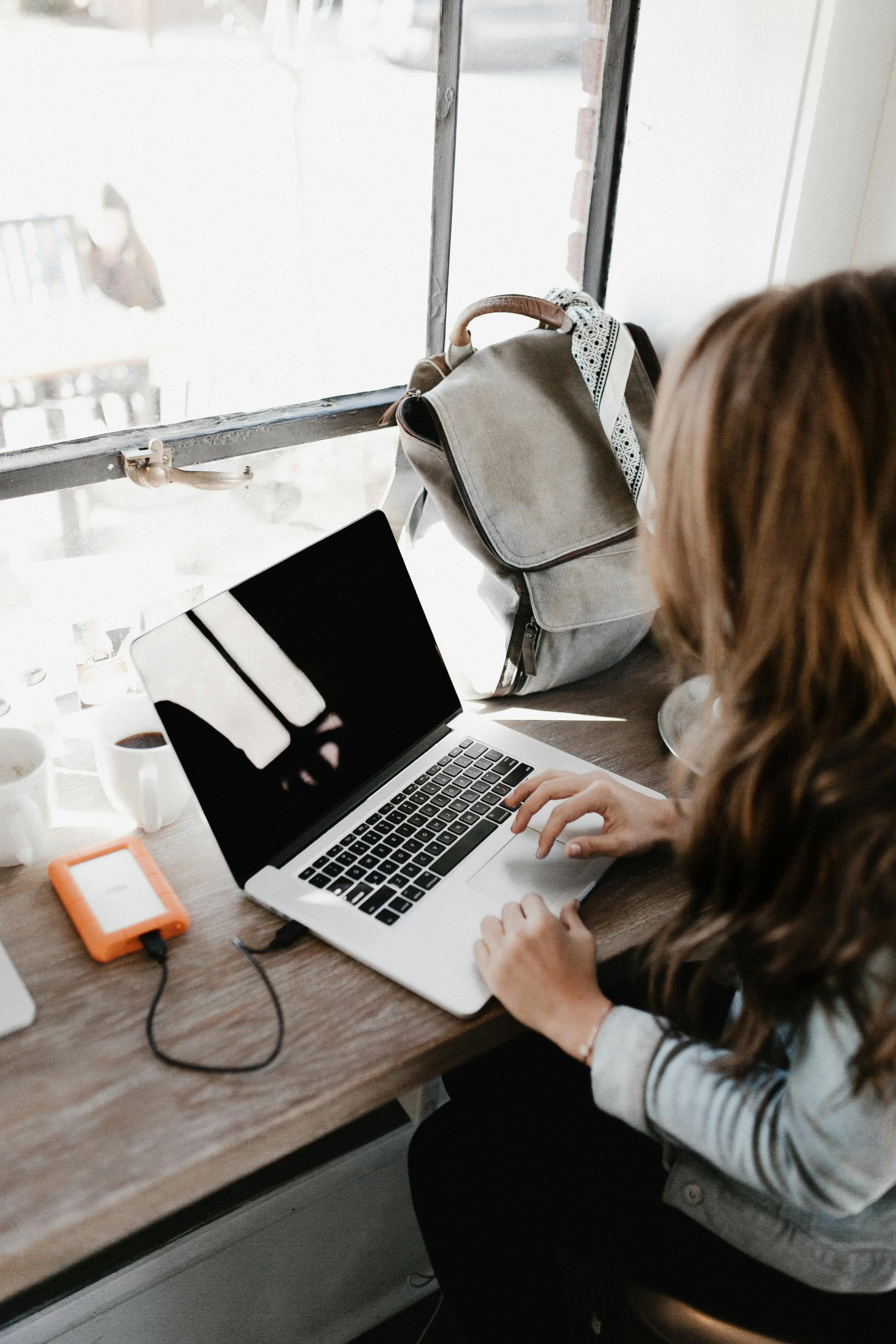 A woman is working on a laptop at a wooden table with some cups of coffee, a small orange device, and a notebook nearby. There is a backpack hanging on a hook on the window frame. She has brown hair, and her right hand is on the keyboard. The window behind her shows an outdoor scene.