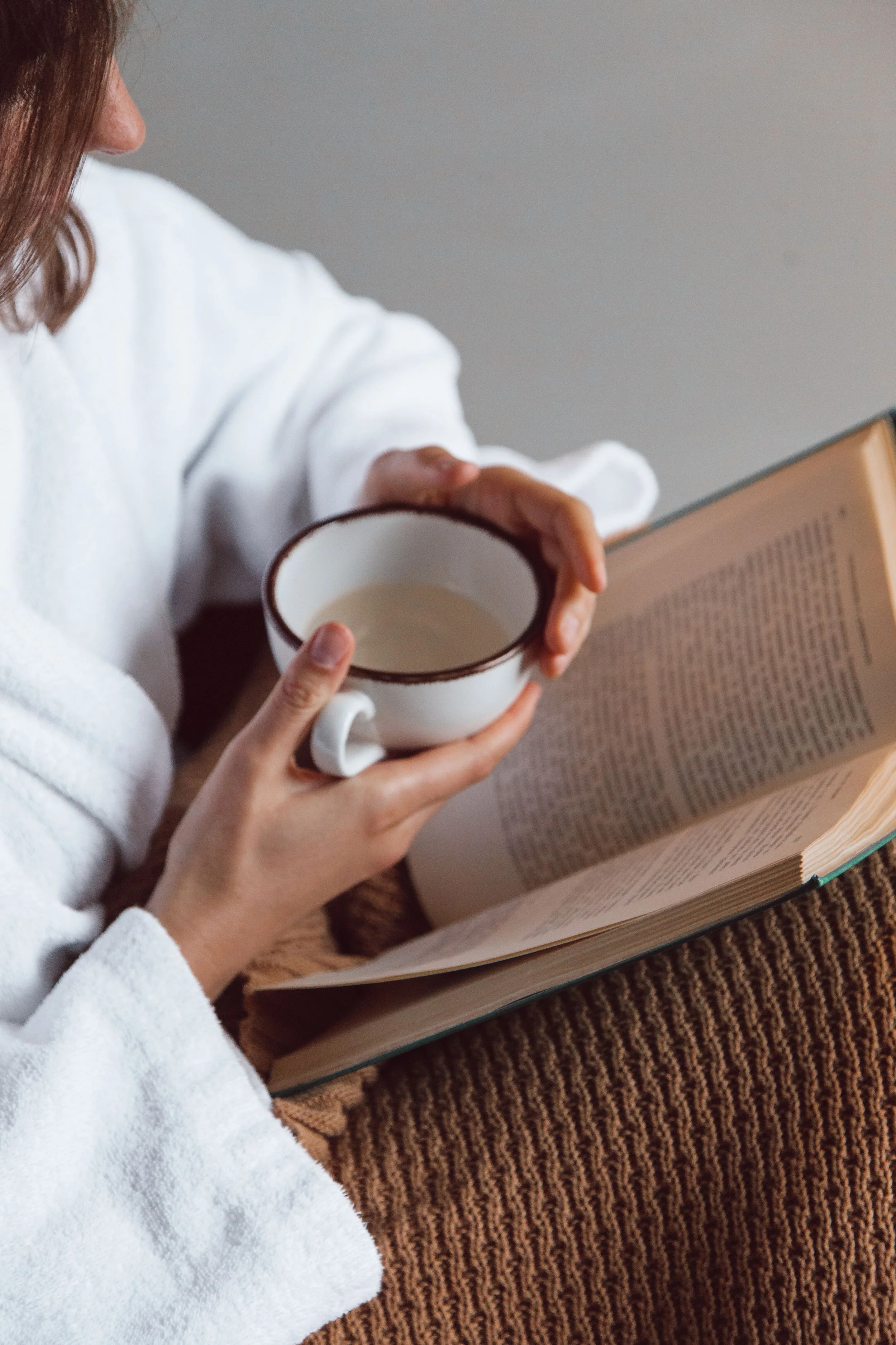 A woman in a white bathrobe sitting on a patterned carpet, holding a cup in one hand and an open book in the other.