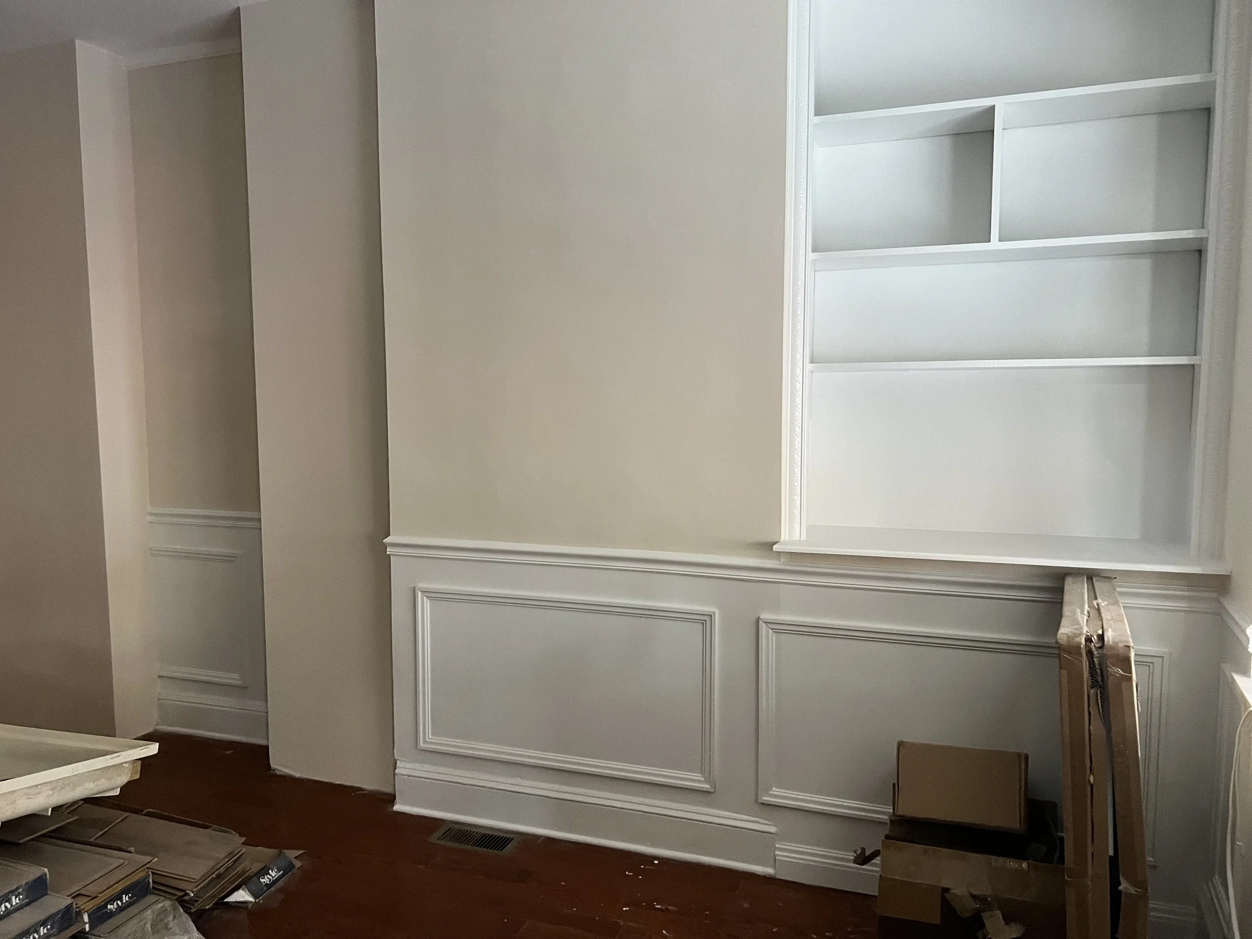 Interior of a room under renovation, showing a built-in white bookshelf and a lower white panel with decorative molding, with boxes and construction materials on the floor.