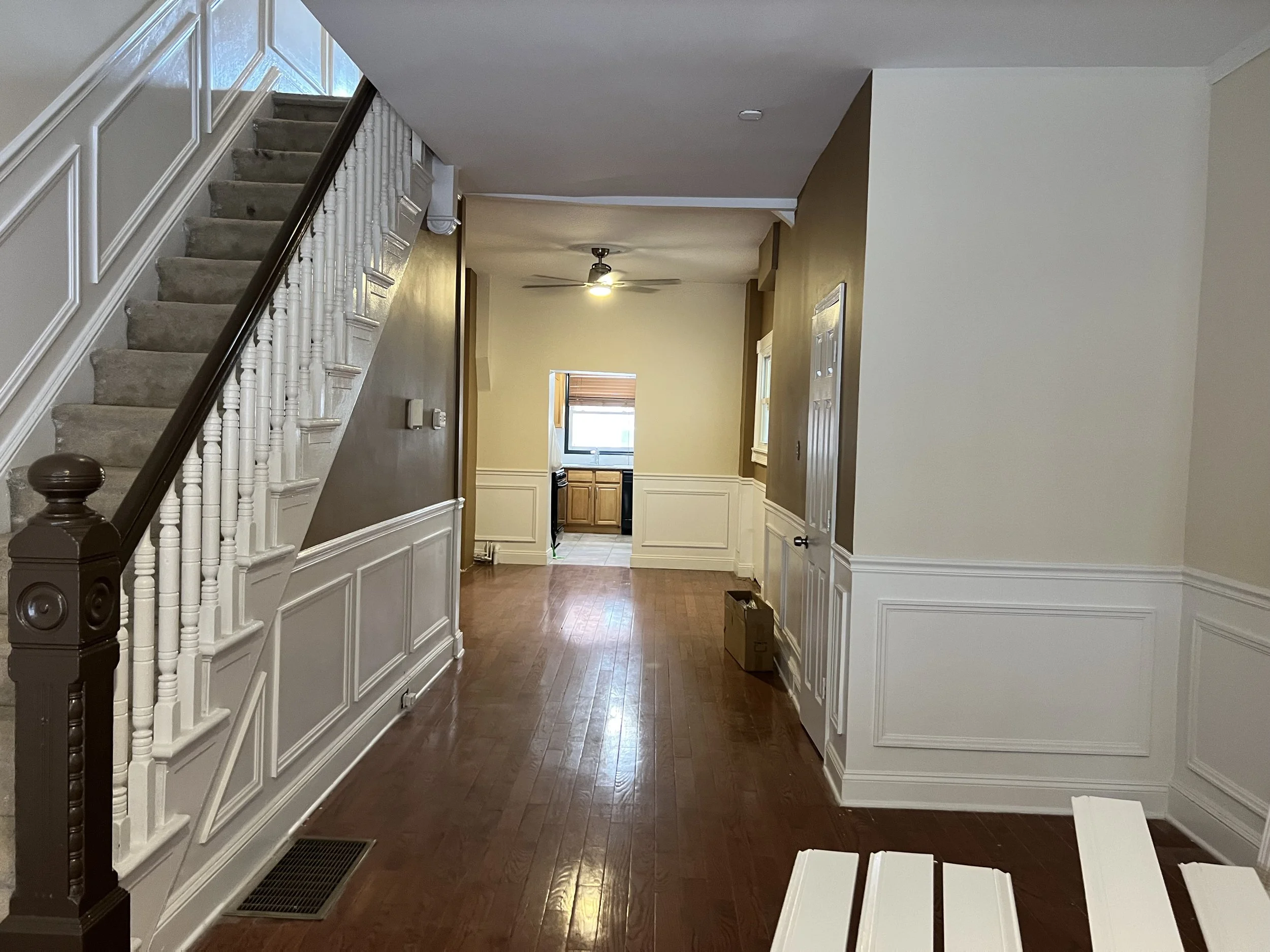 Interior of a home with a staircase on the left, wood flooring, and a kitchen in the background with a ceiling fan and window. Walls are painted in beige and white wainscoting decorates the lower half.