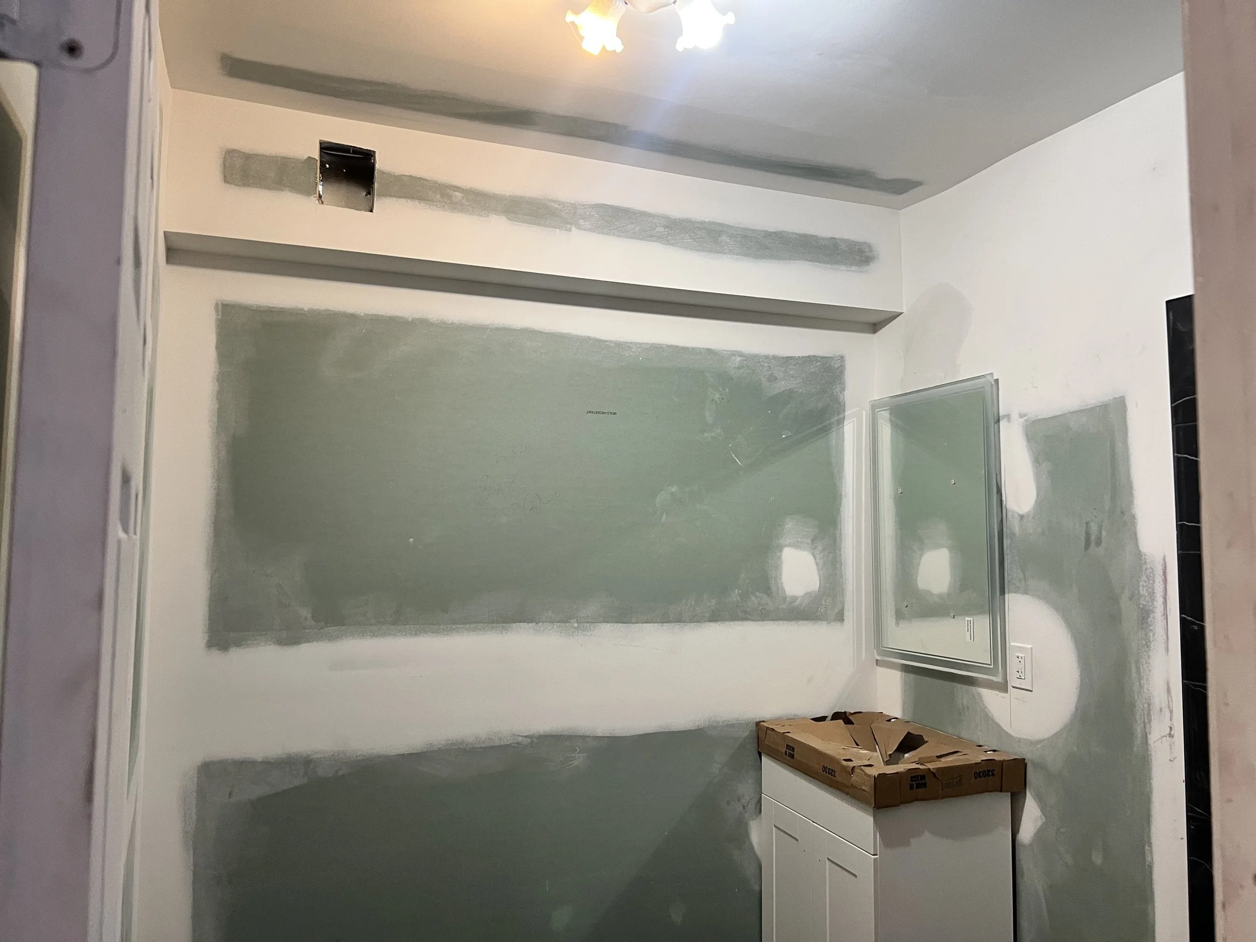 A room under construction with partially painted walls, a small white cabinet with a cardboard box on top, a window with a plastic frame, and a ceiling light fixture.