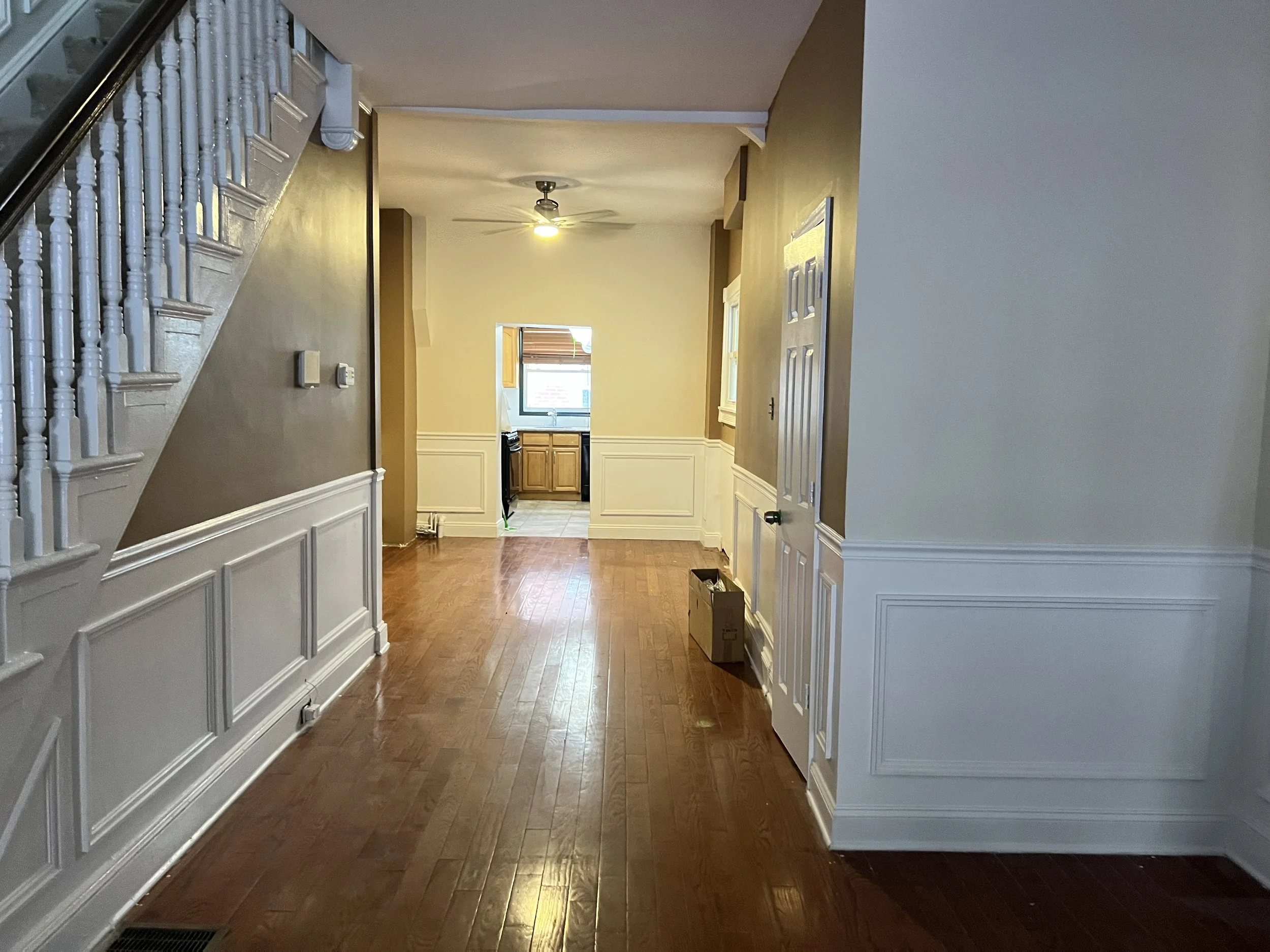Interior view of a house hallway leading to a kitchen, with hardwood floors, white wainscoting, beige walls, staircase on the left, and a ceiling fan with a light on