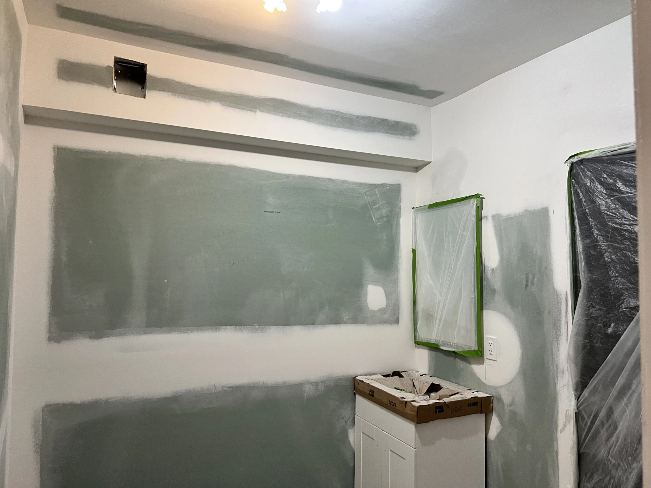 A room undergoing renovation with walls painted in an unfinished color, covered with patchy areas of green paint. There is a small cabinet covered with a cardboard box, pushed against the wall. A plastic-covered window is taped with green painter's t