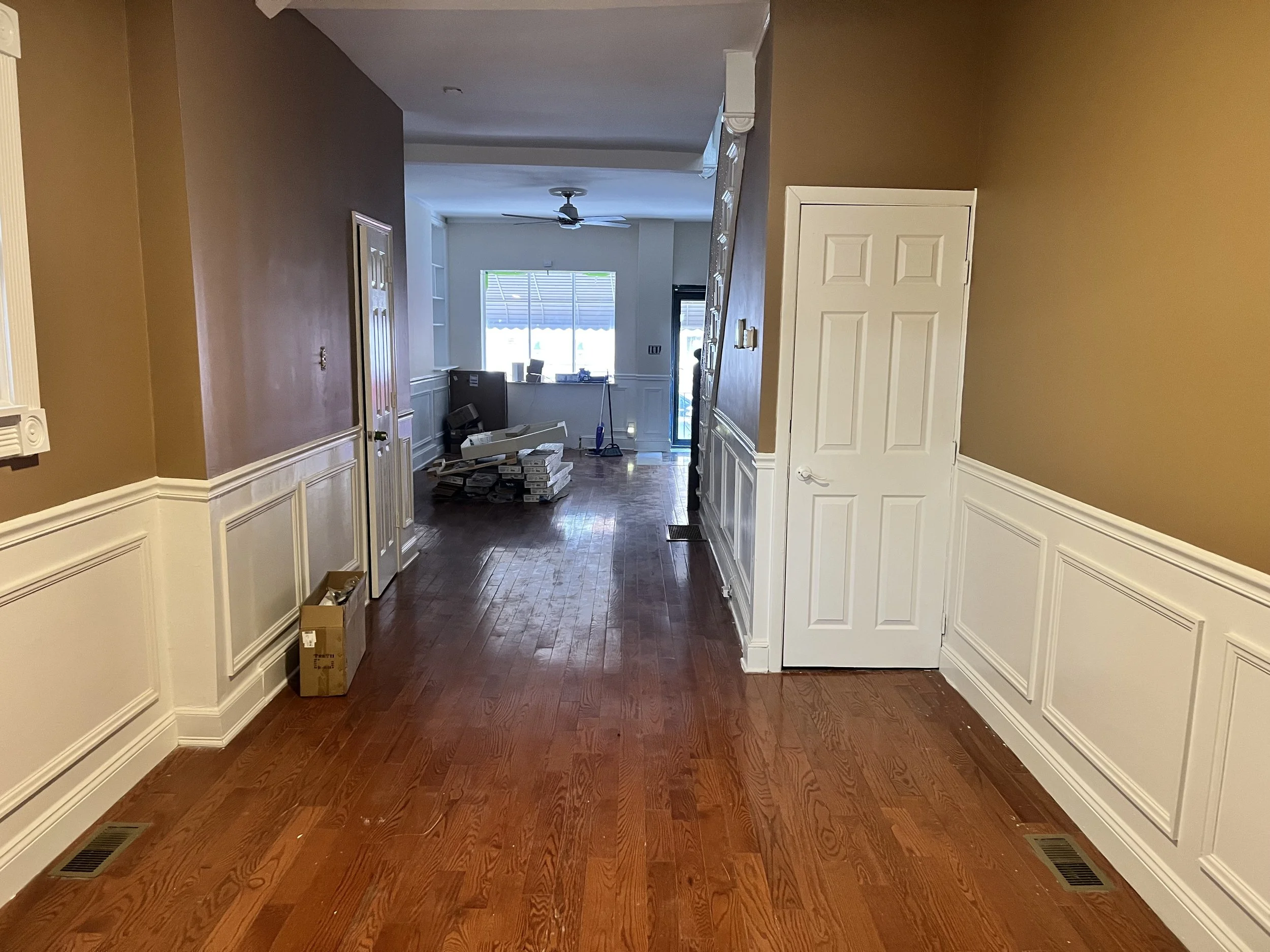 Interior view of a house under renovation, with hardwood floors, partially filled boxes, and construction materials near a window and doorway.