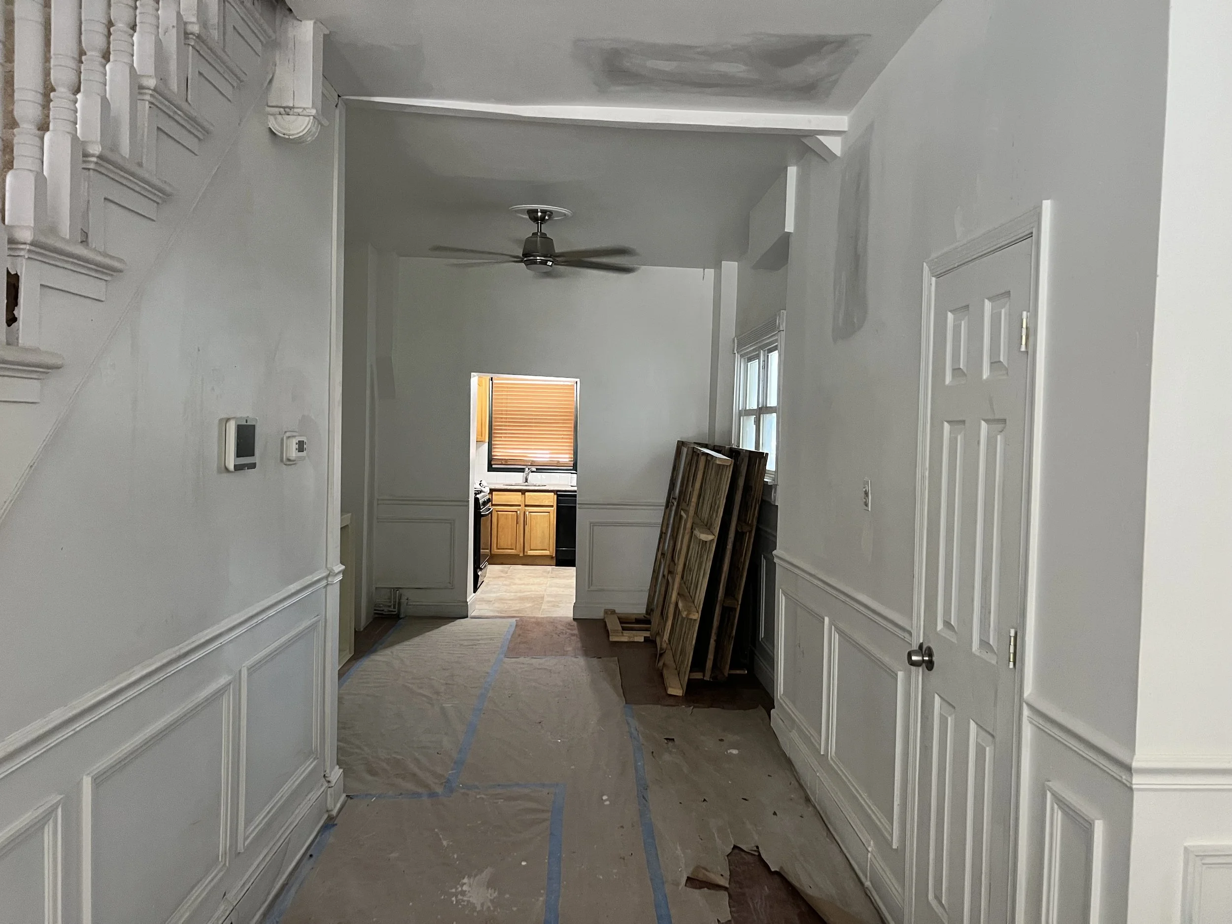 Interior of a house under renovation with white walls, a staircase on the left, wooden cabinets visible through an opening, and painting or drywall work in progress on the walls and ceiling.