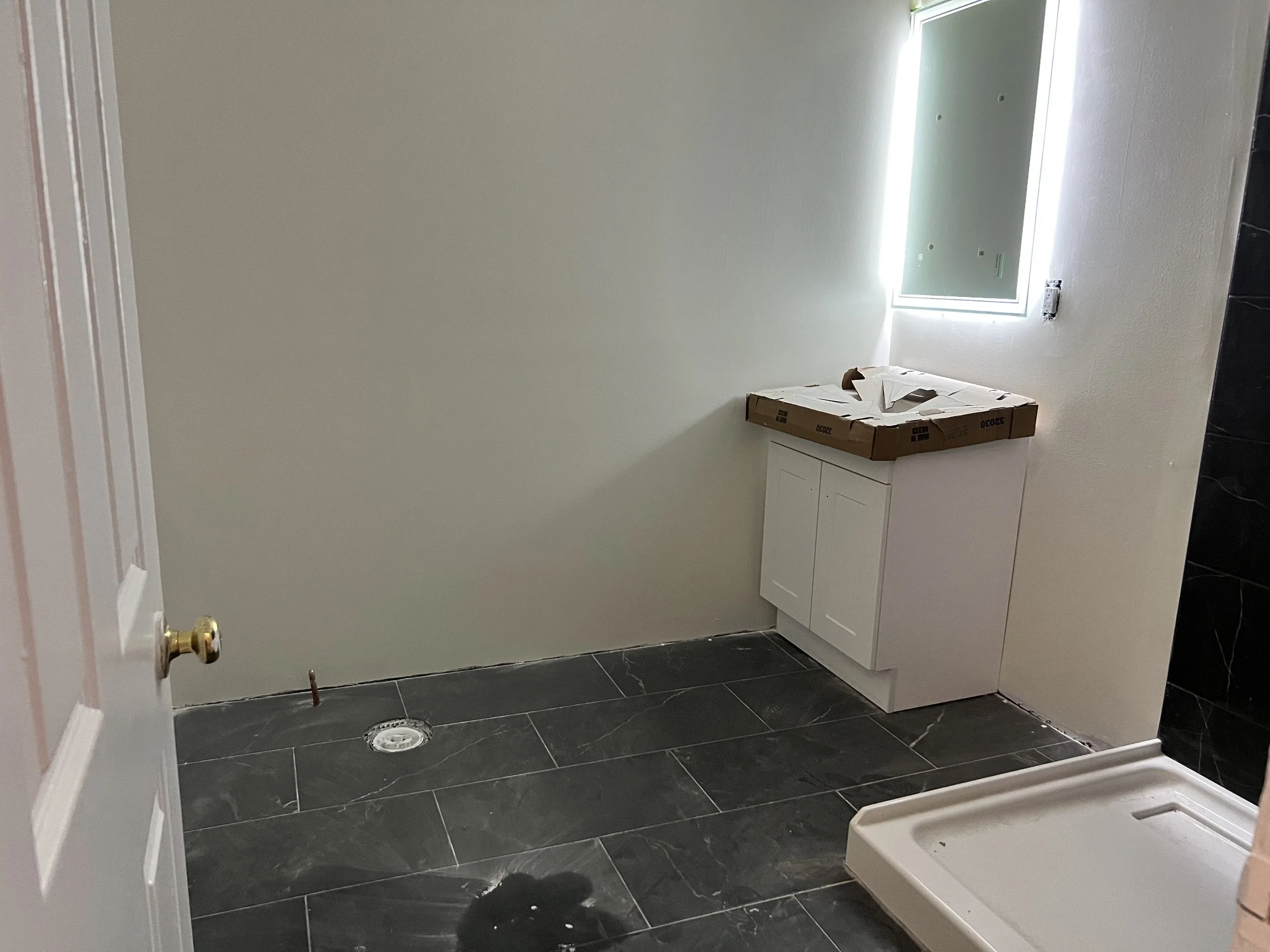 Empty bathroom with black marble floor tiles, a small white cabinet, a window, and a shower base in the corner, with construction materials on the cabinet.
