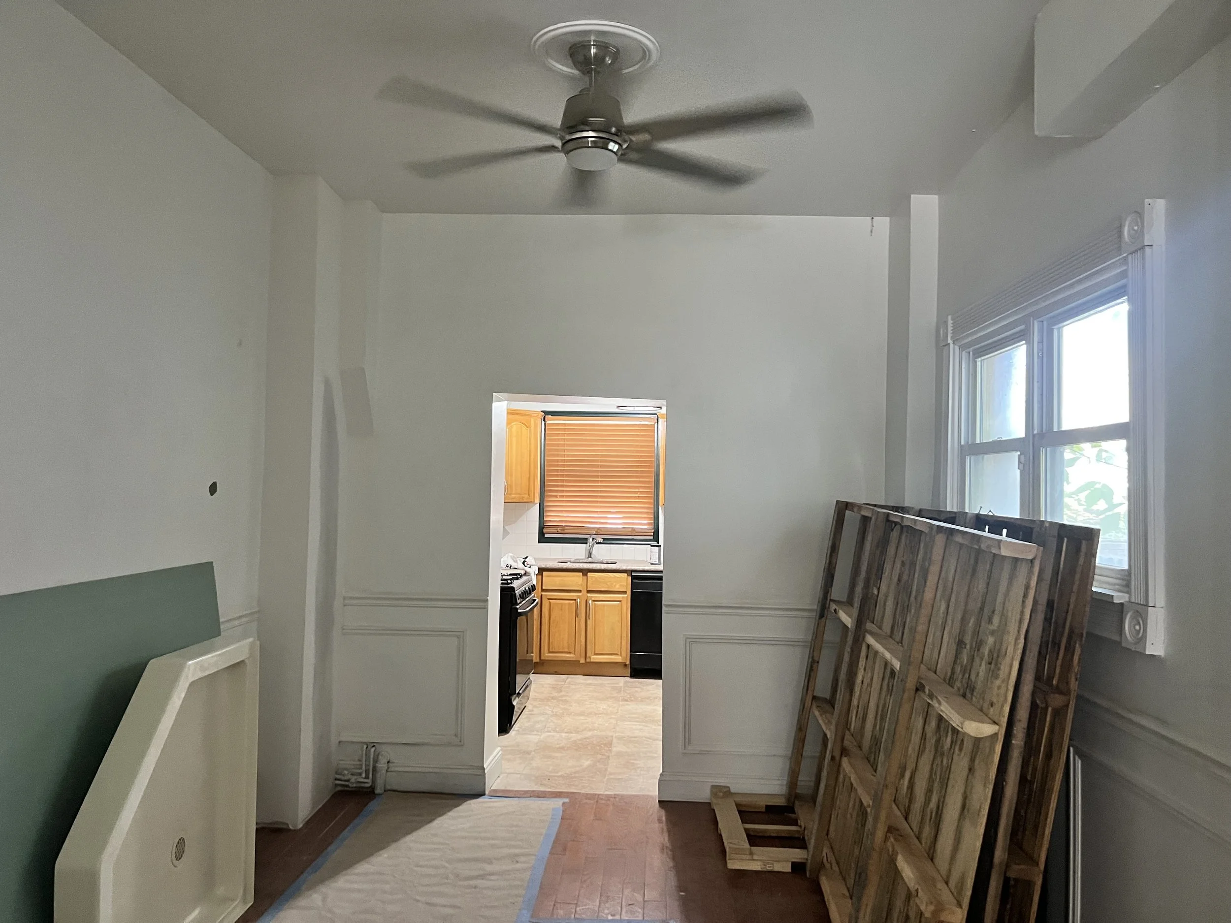 Empty room with a ceiling fan, large window, and a doorway leading to a kitchen with wooden cabinets and a window with blinds. Various wooden pallets and paneling are leaning against the wall.
