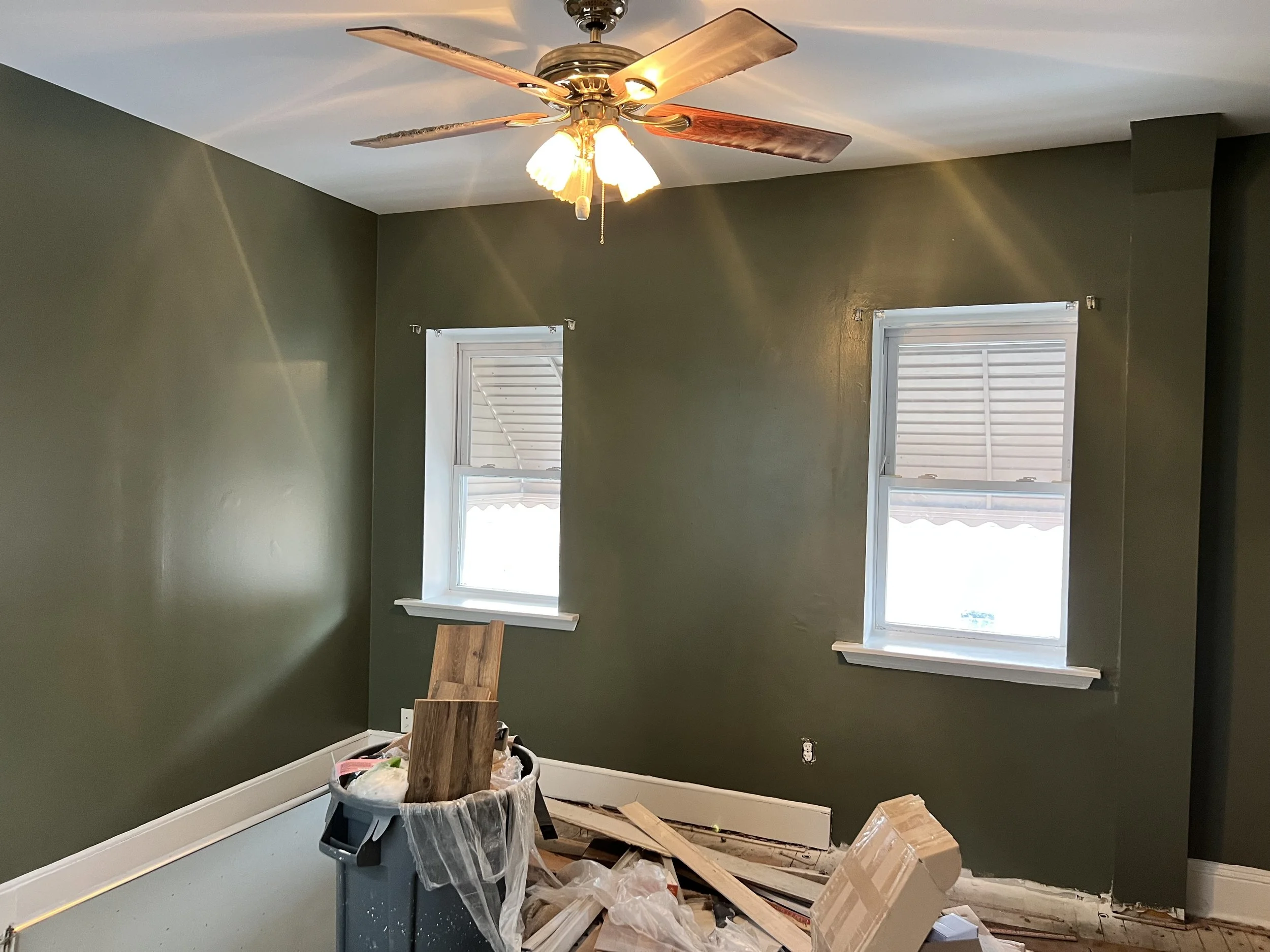 Room with two windows, green walls, ceiling fan with lights, and renovation debris on the floor, including wood scraps and a trash bin