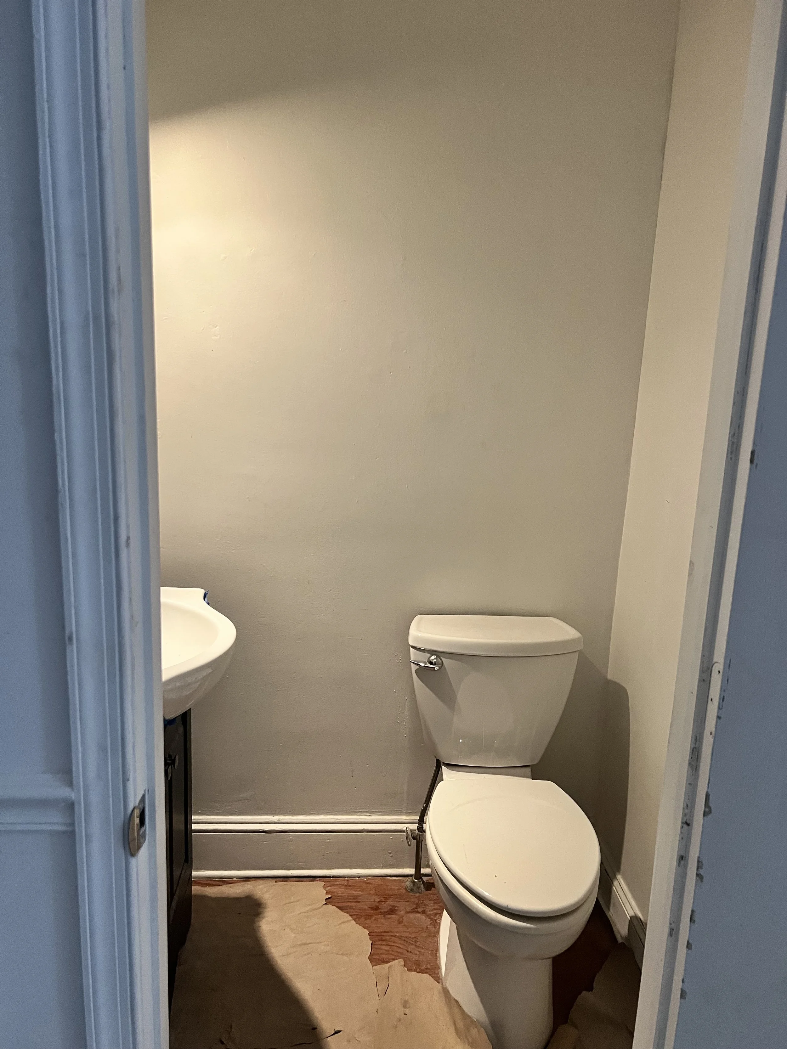A small bathroom with a white toilet, a partial view of a white sink on a dark cabinet, light-colored walls, and a wooden floor.