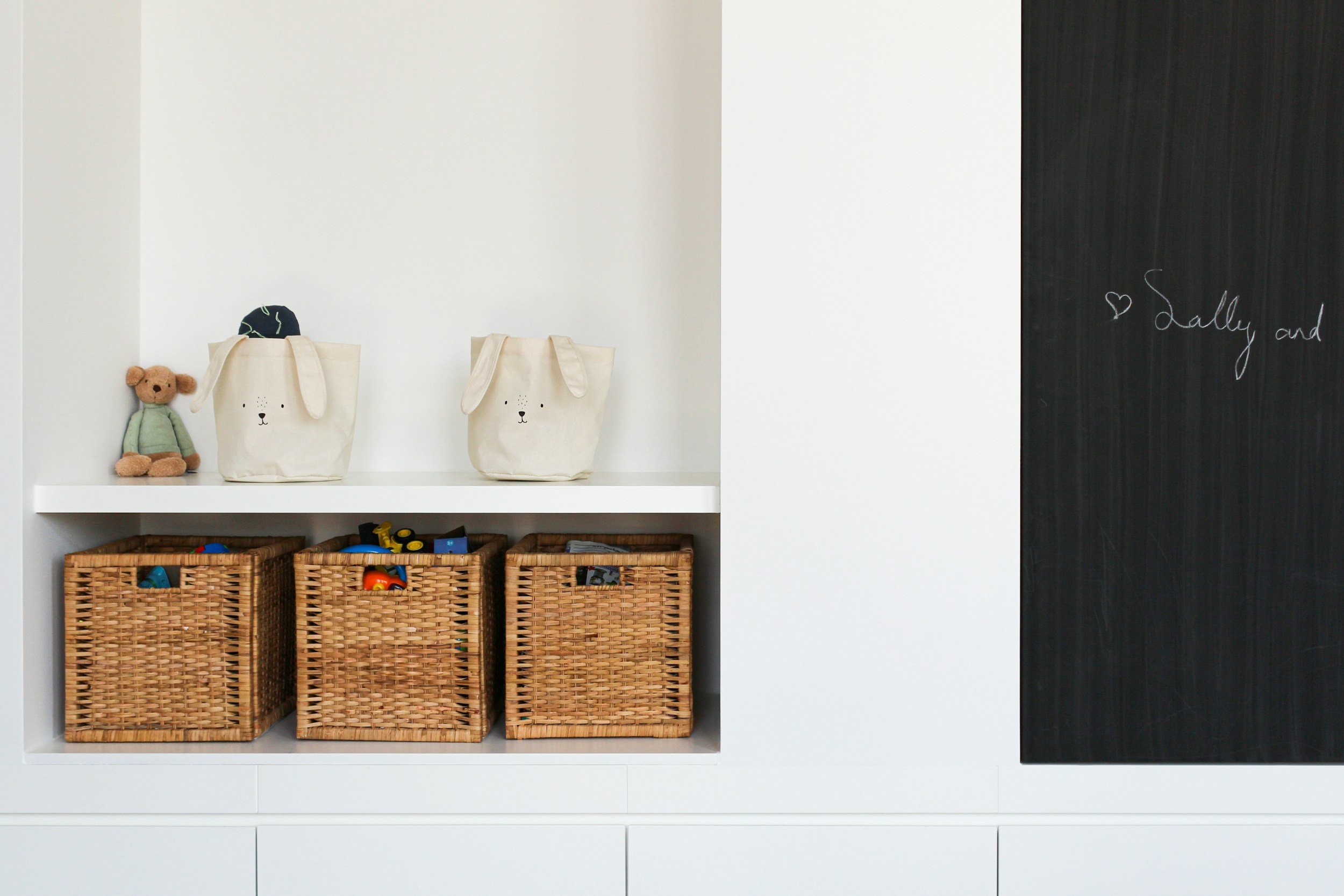 Storage area with two animal face bags on a shelf, a small teddy bear, three woven baskets filled with toys, and a blackboard with writing that says 'Sally and'.