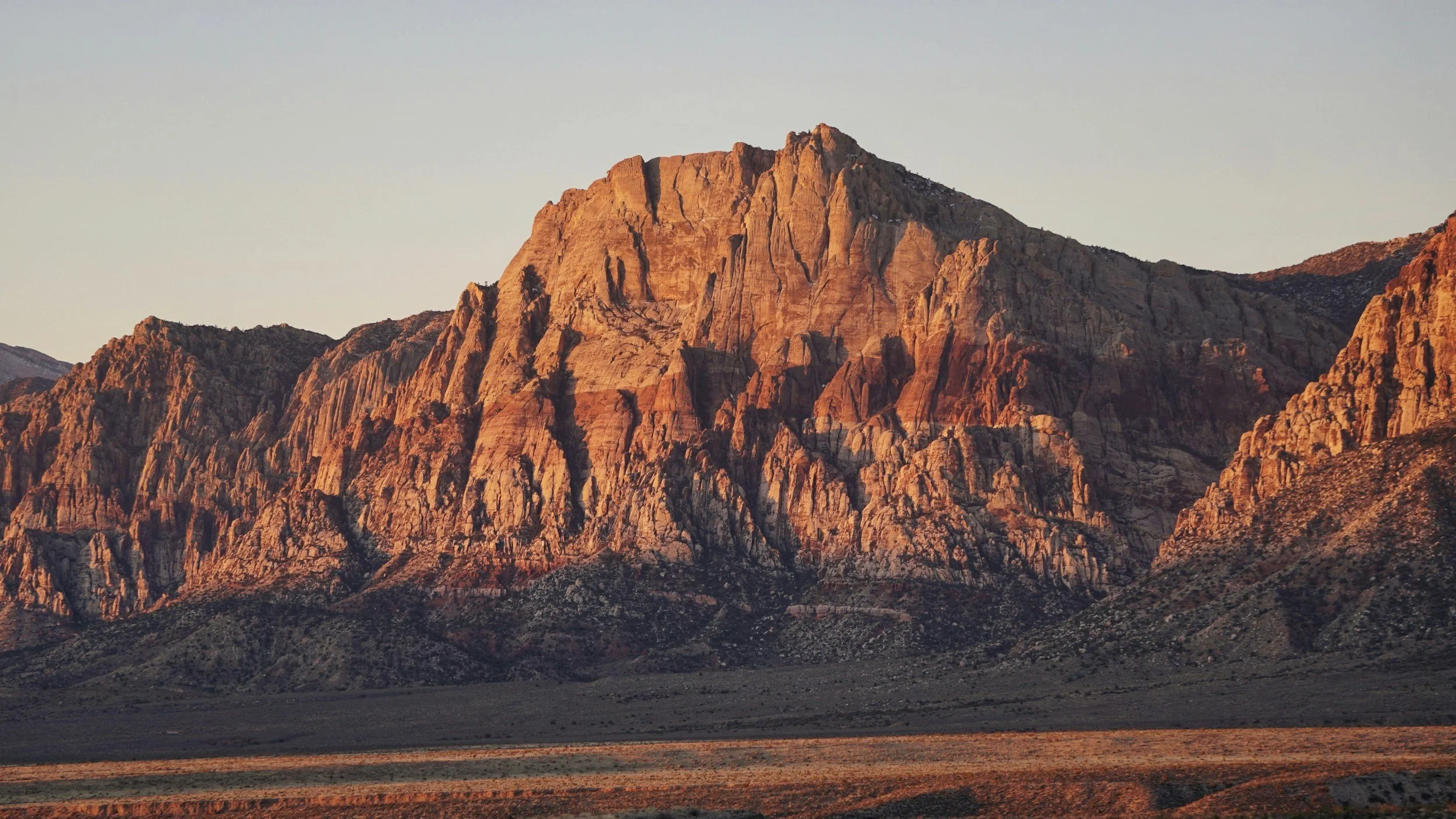 Sunset illuminating rugged desert mountain landscape with reddish and tan rocky formations.