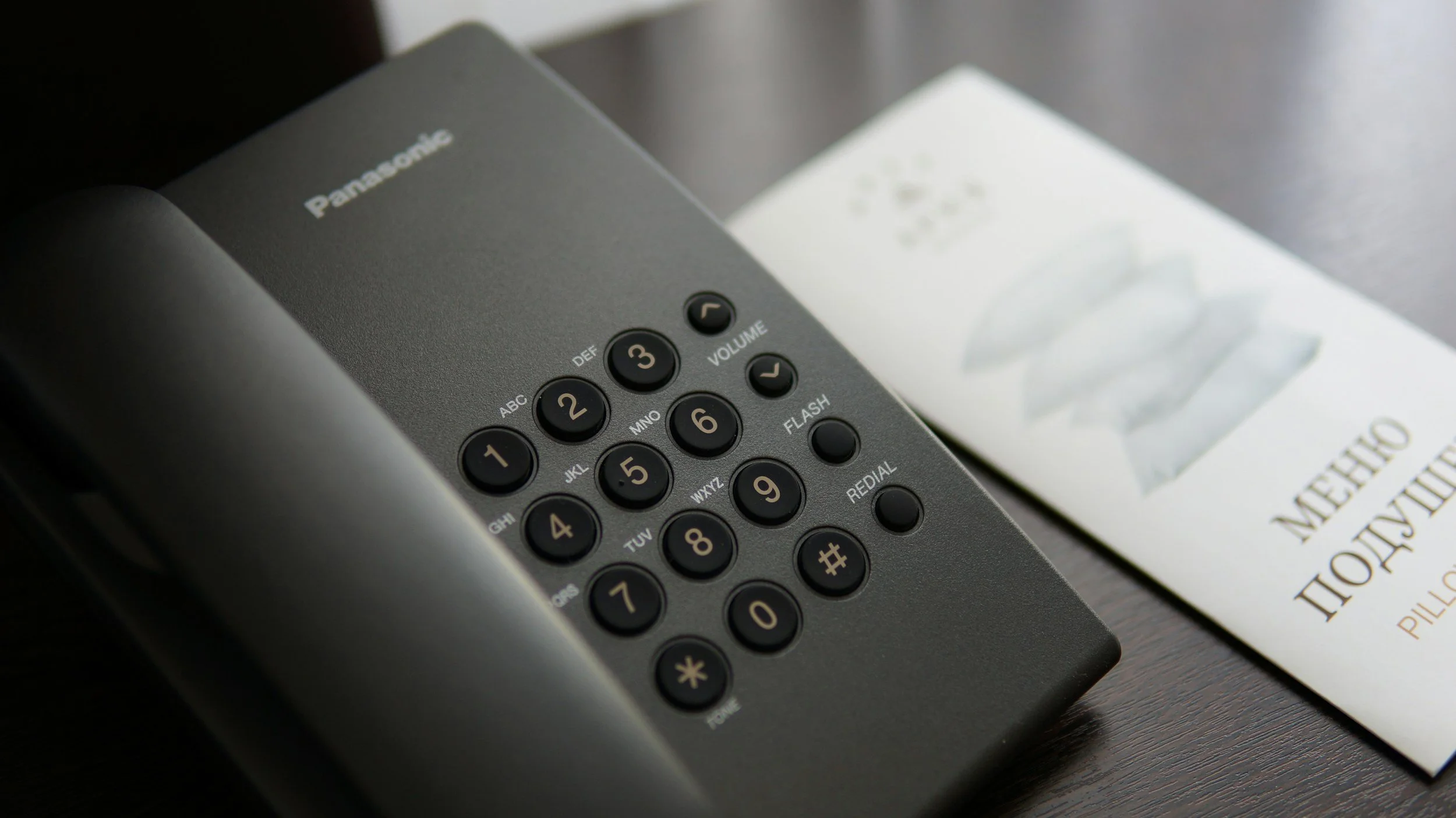 Close-up of a Panasonic landline phone with black buttons on a dark surface, alongside a pamphlet titled 'MEET THE PILLOW'.