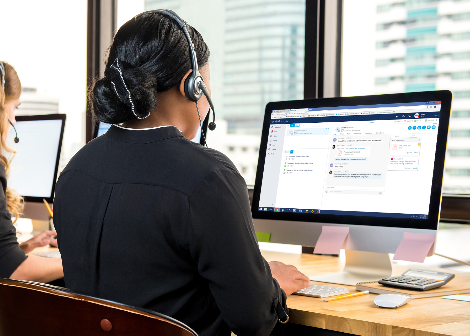 A woman in a black shirt wearing a headset, working at a computer station in an office, with another woman in the background also working on a computer.