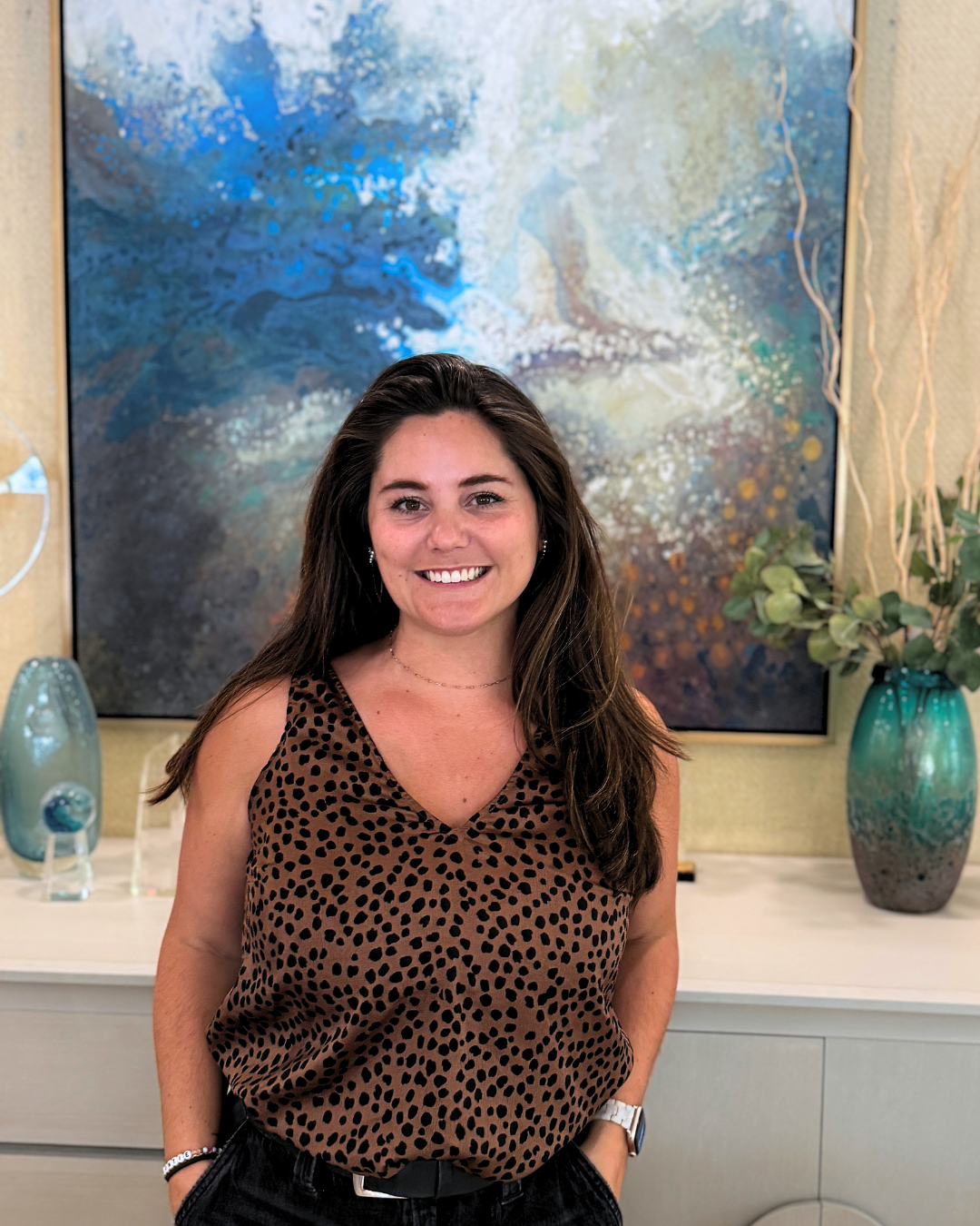 A smiling woman with long brown hair standing in front of an abstract painting with blue, white, and beige tones, and decorative vases with green plants.