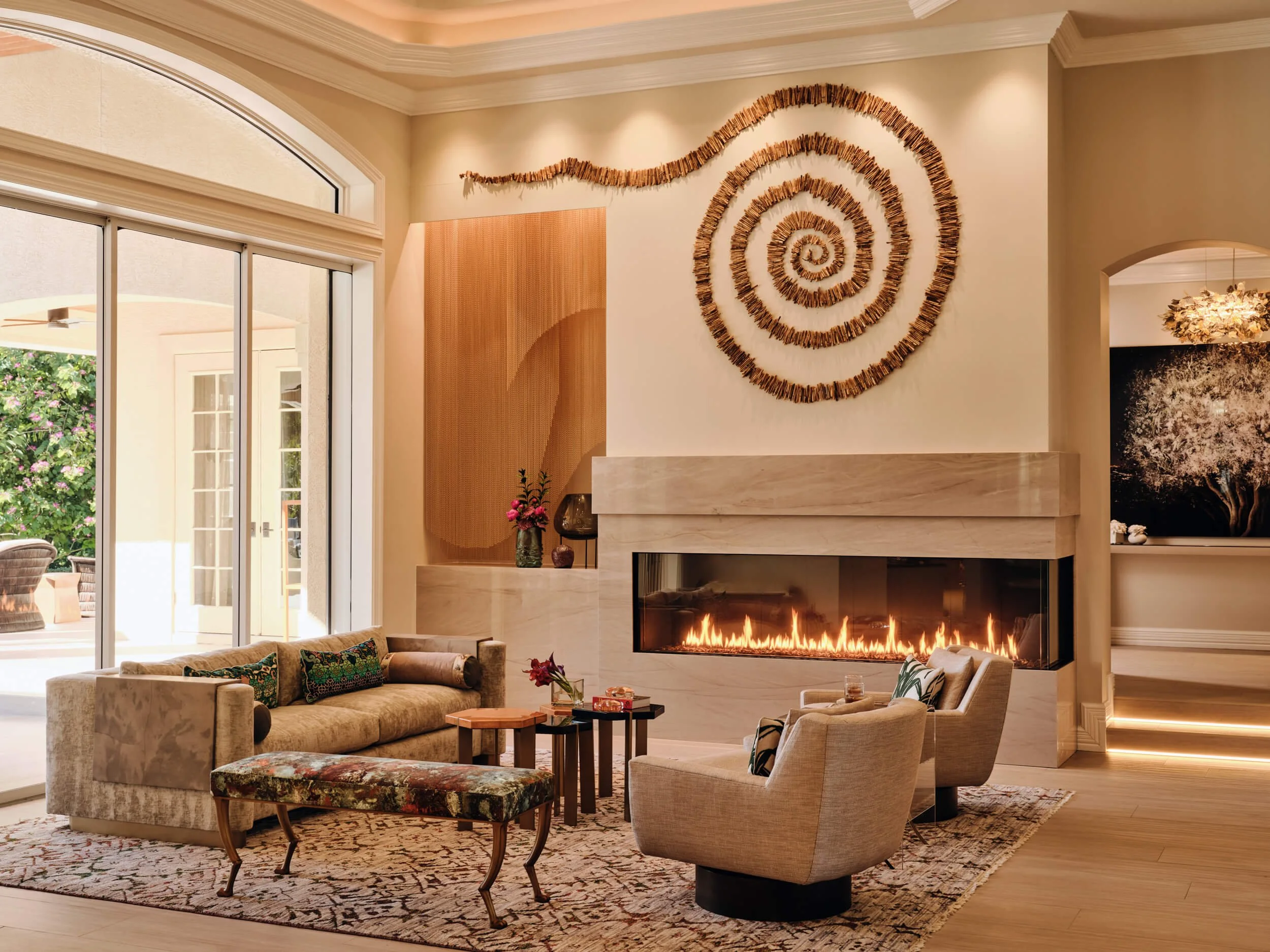 A cozy living room with a fireplace, decorated with spiral wood wall art, and a seating area with a sofa and armchairs. Large sliding glass doors lead to an outdoor patio with greenery and patio furniture.