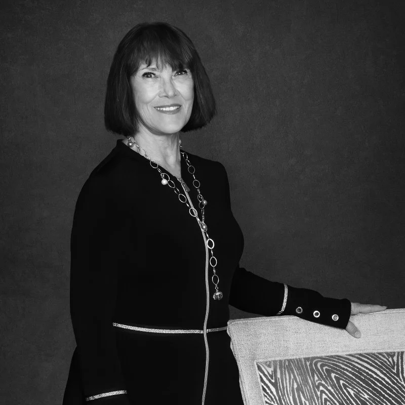 A woman with dark hair, smiling, wearing a black dress with decorative zipper and rings, and a long necklace. She is standing against a dark background, touching a patterned chair.