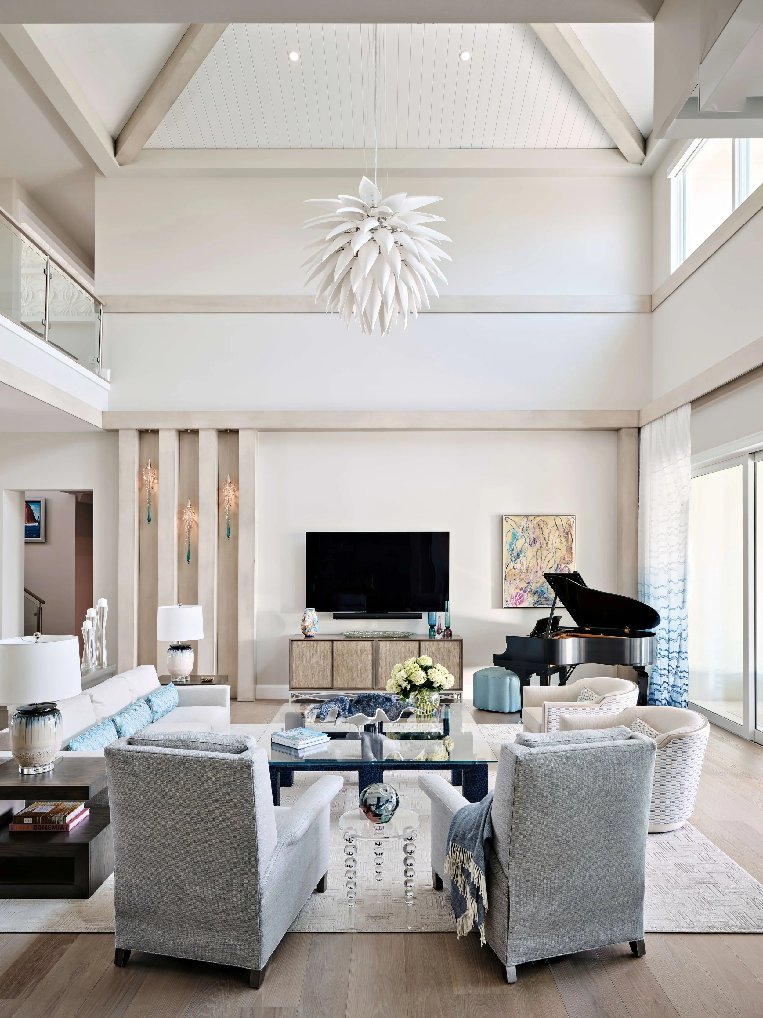 Bright living room with high vaulted ceiling, white walls, hanging chandelier, large flat-screen TV, grand piano, and contemporary furniture including armchairs, coffee tables, and decorative vases.