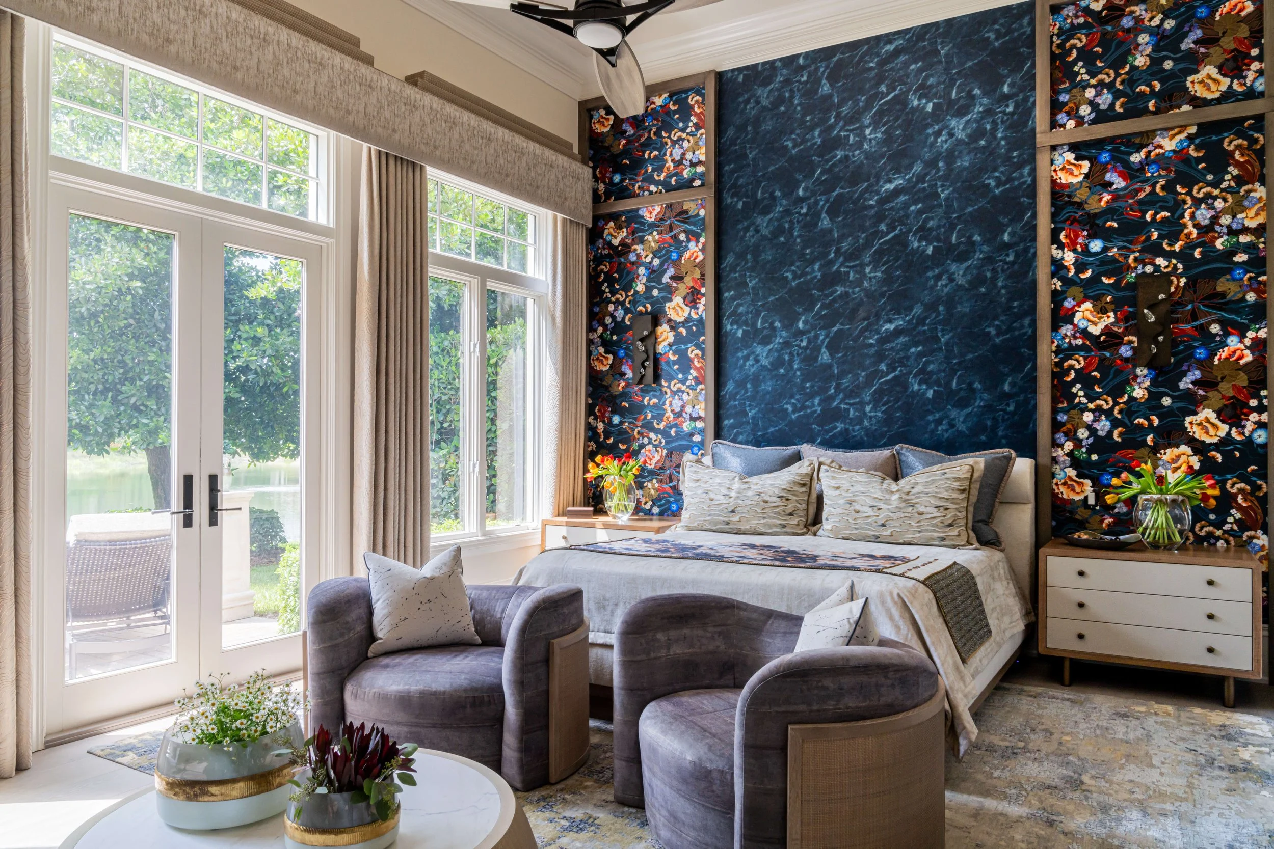 Living room with large windows, floral wallpapered accent wall, beige curtains, a beige bed with pillows, a gray velvet sofa, a round white coffee table with potted plants, and a ceiling fan.