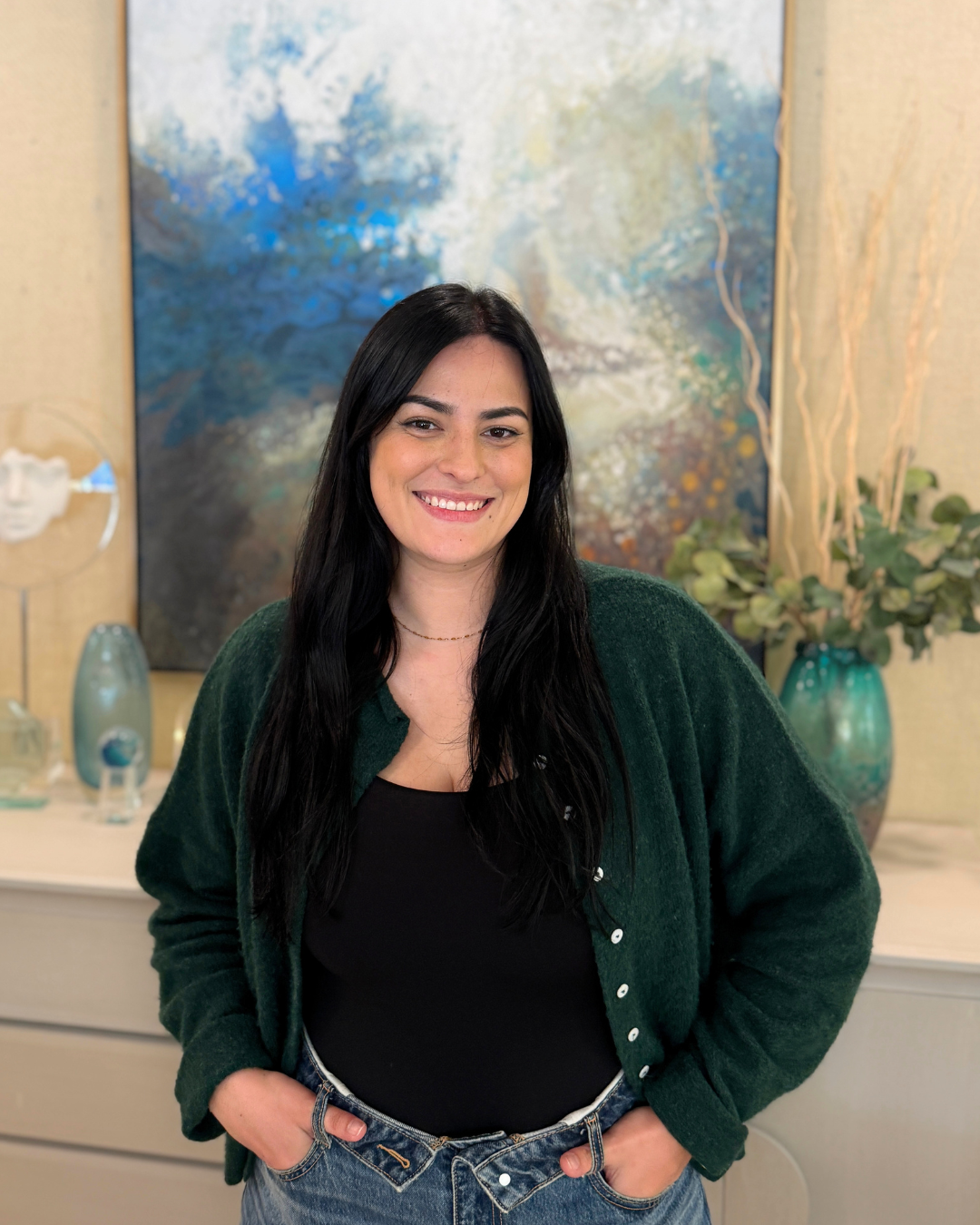 A woman with long dark hair, wearing a green cardigan over a black top and jeans, smiling while standing in front of an abstract painting with blue, green, and beige tones, and a decorated background with vases and plants.
