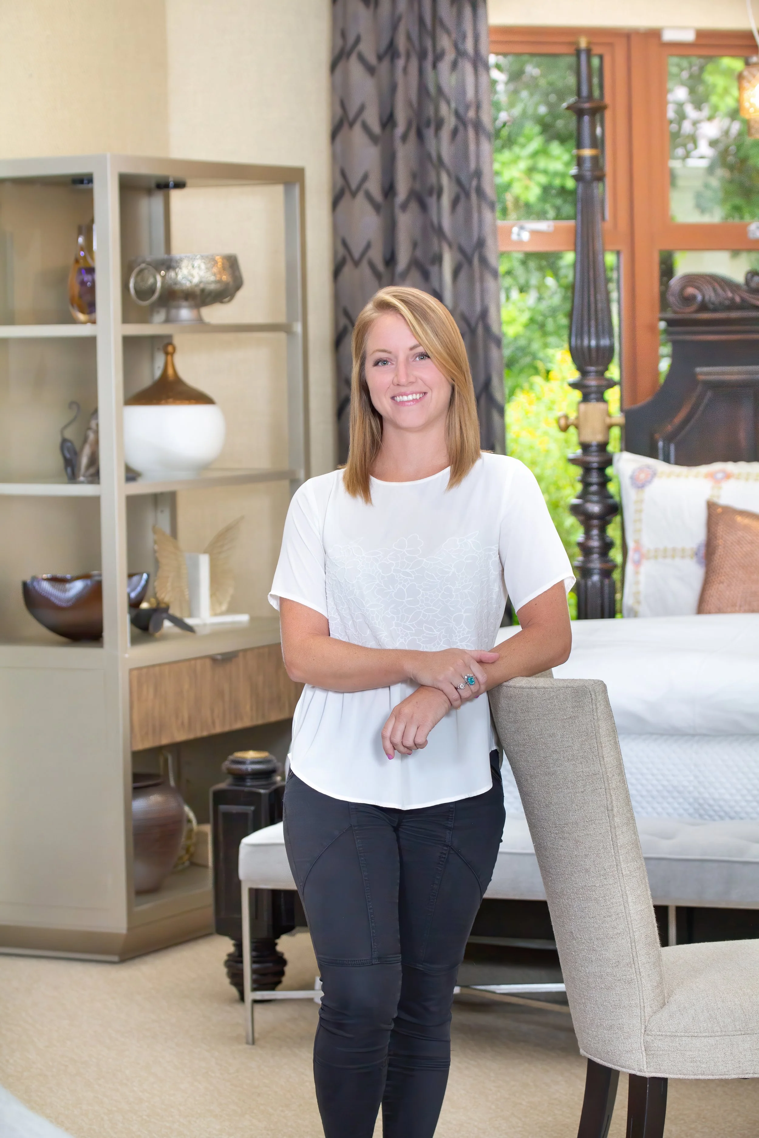 A smiling woman with shoulder-length blonde hair, wearing a white blouse and black pants, standing in a well-decorated living room near a beige armchair and a shelf with decorative items, with a large window and outdoor greenery in the background.