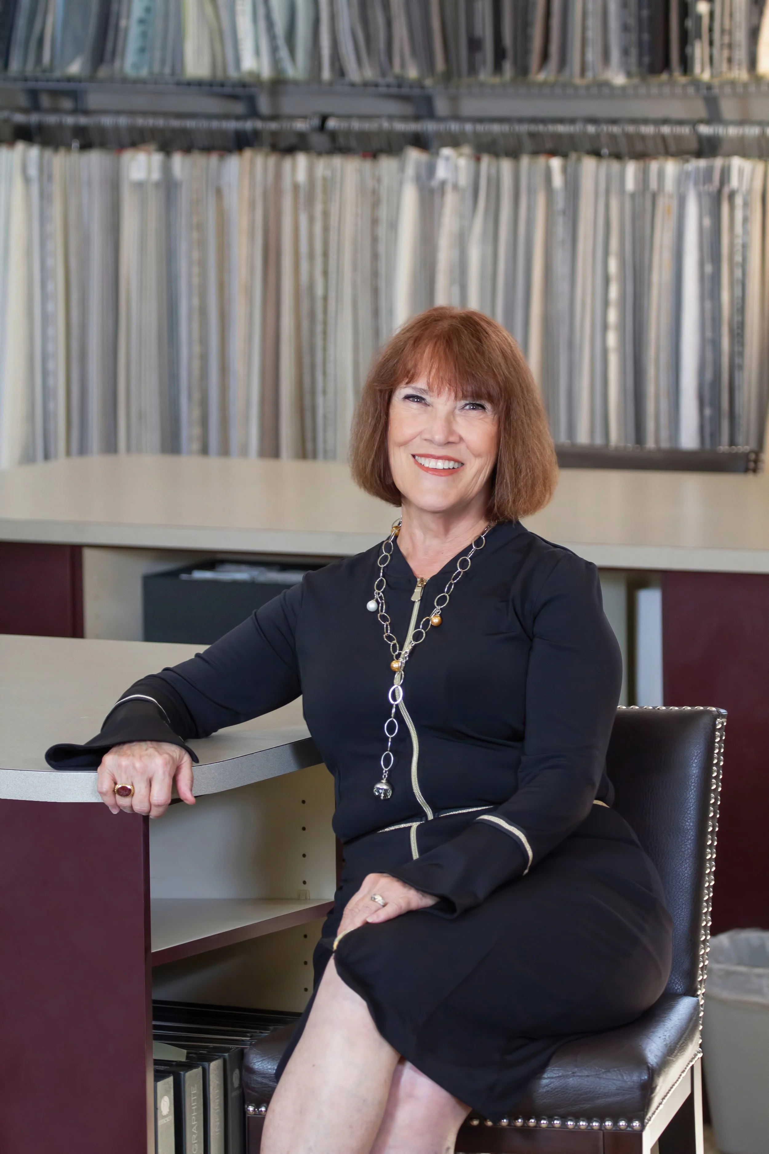 A smiling middle-aged woman with shoulder-length reddish hair, wearing a black dress with a zipper and a long necklace, sitting at a desk in an office, with a background of bookshelves filled with binders and books.